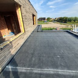 Usługi dekarskie Kamil Kowalski - Płaski dach pokryty czarną membraną bitumiczną, widoczne łączenia, fragment muru z cegły klinkierowej i niezabudowane otwory okienne, w tle zabudowa jednorodzinna.