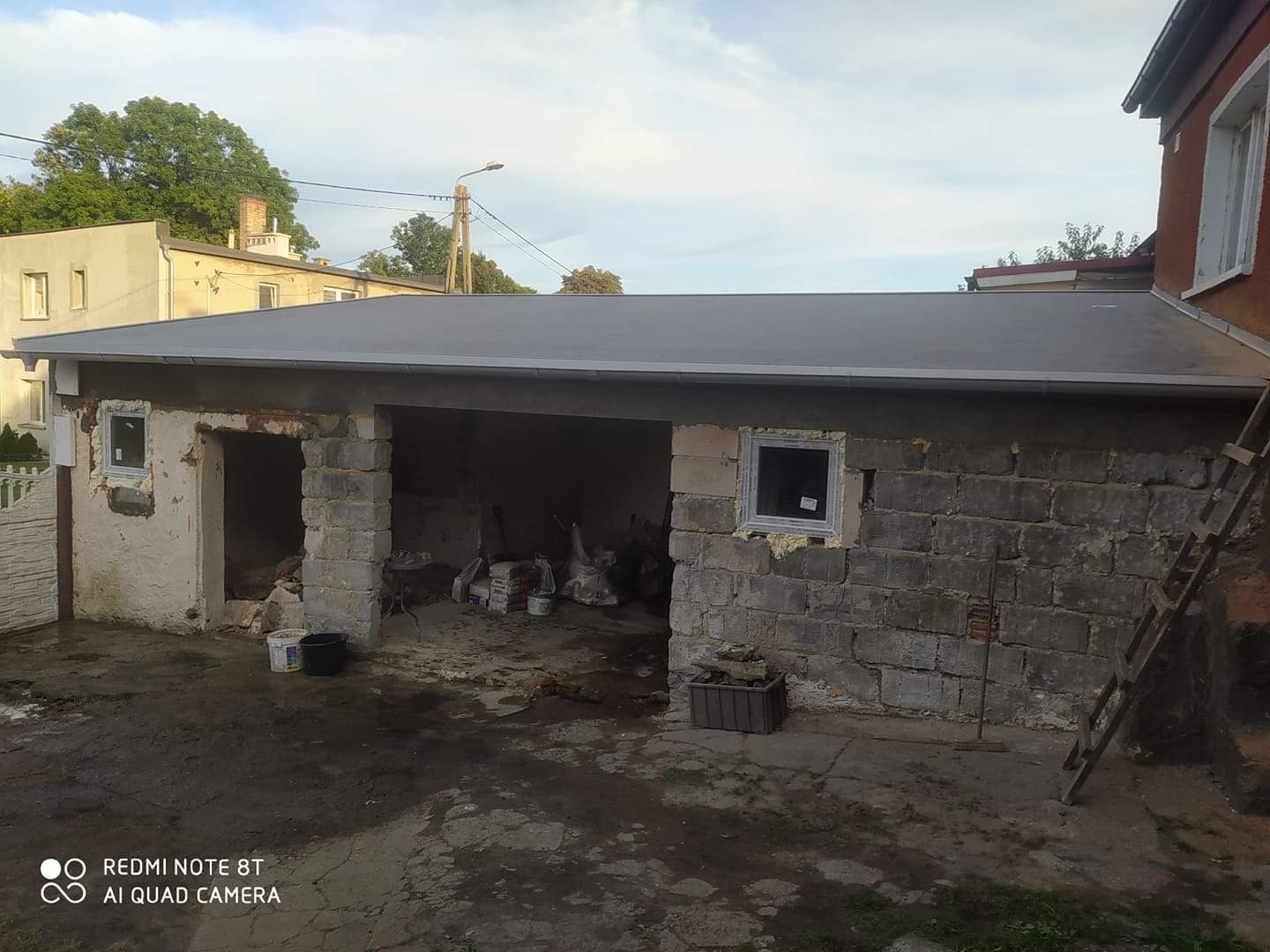 Nisko położony, płaski dach pokryty papą na murowanym garażu w trakcie remontu, z widoczną drabiną opartą o ścianę i oknem z pianką montażową.