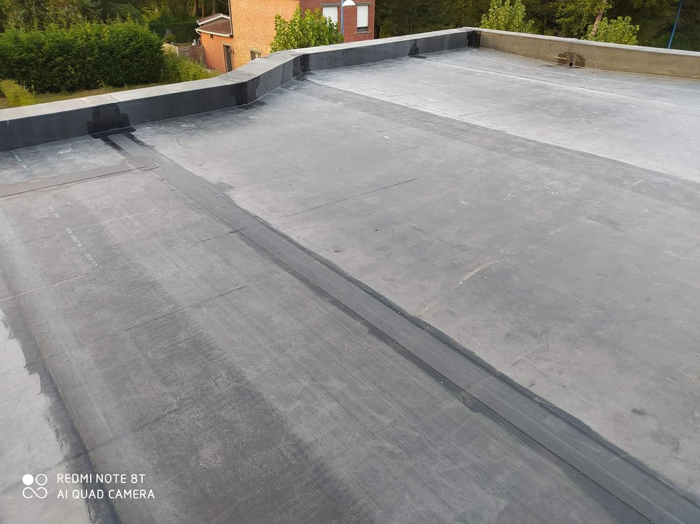 Płaski dach pokryty papą bitumiczną z widocznymi łączeniami i obróbkami blacharskimi, w tle budynek mieszkalny i zieleń.