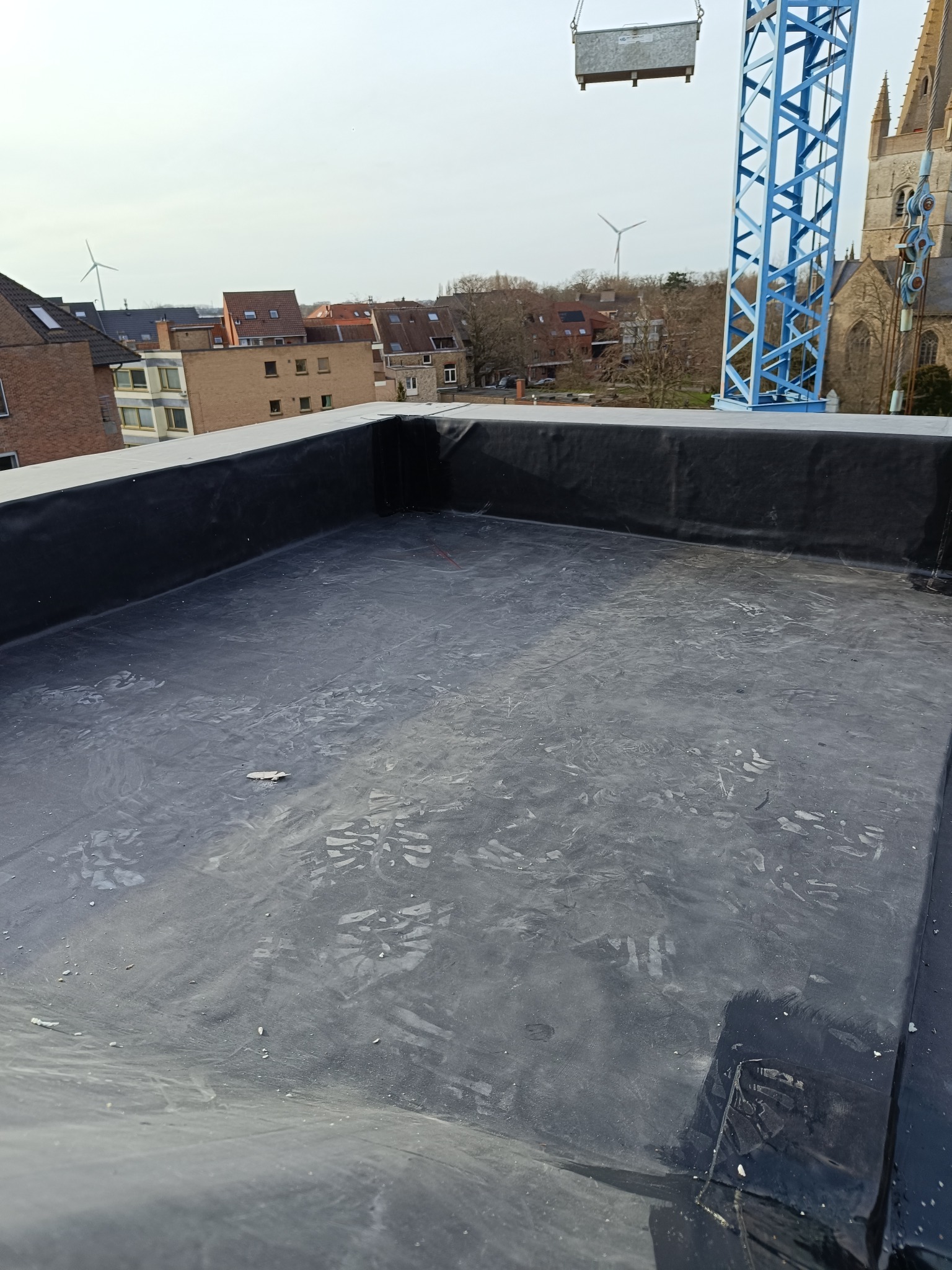 Płaski dach pokryty czarną membraną, widoczne ślady butów, w tle zabudowa miejska z kościołem i wiatrakami, niebieski dźwig w prawym górnym rogu.