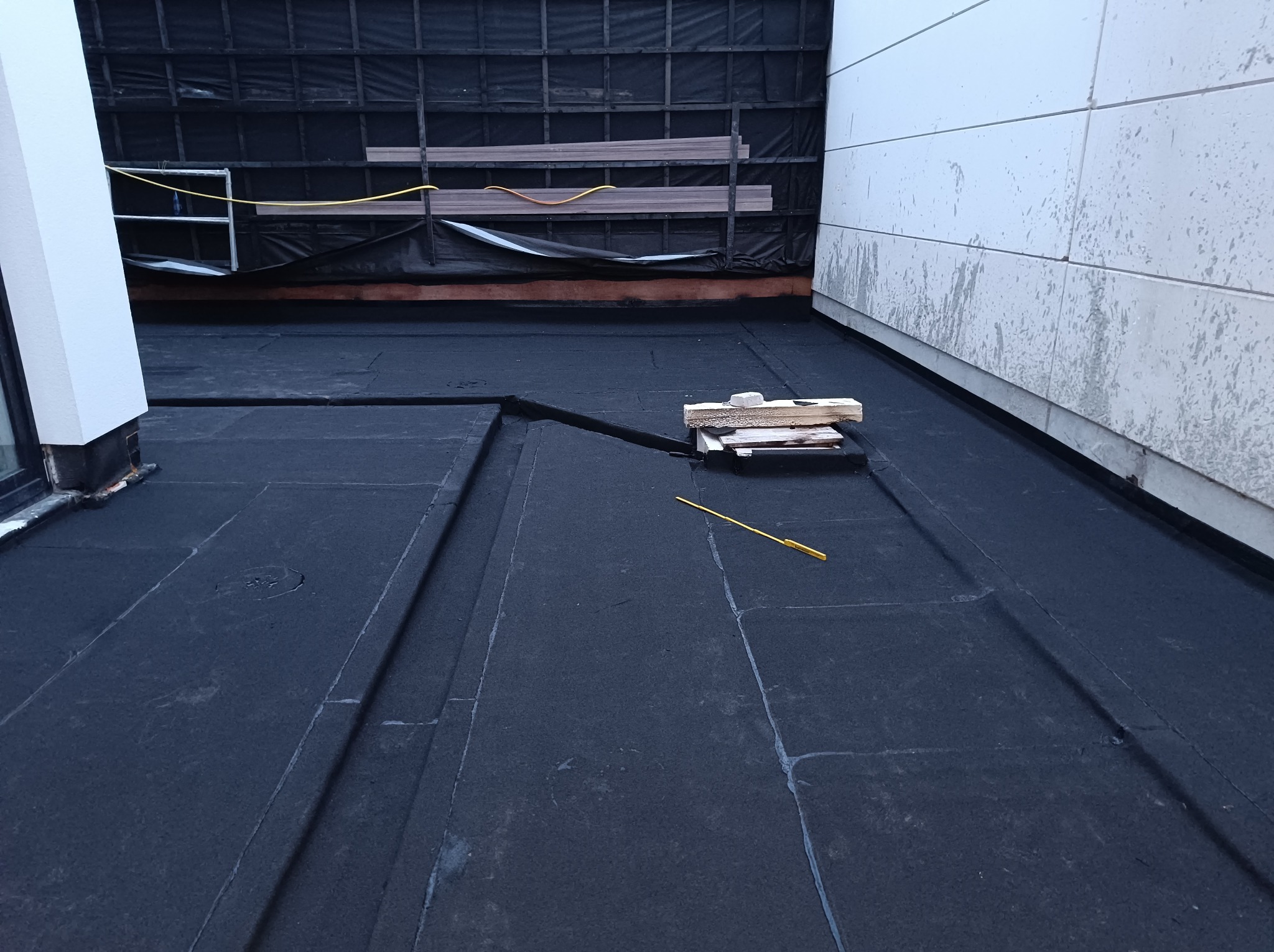 Czarna papa na płaskim dachu w Brodnicy, widoczne łączenia, na górze zaplecze budowy z deskami i rusztowaniem, po prawej biała ściana budynku.