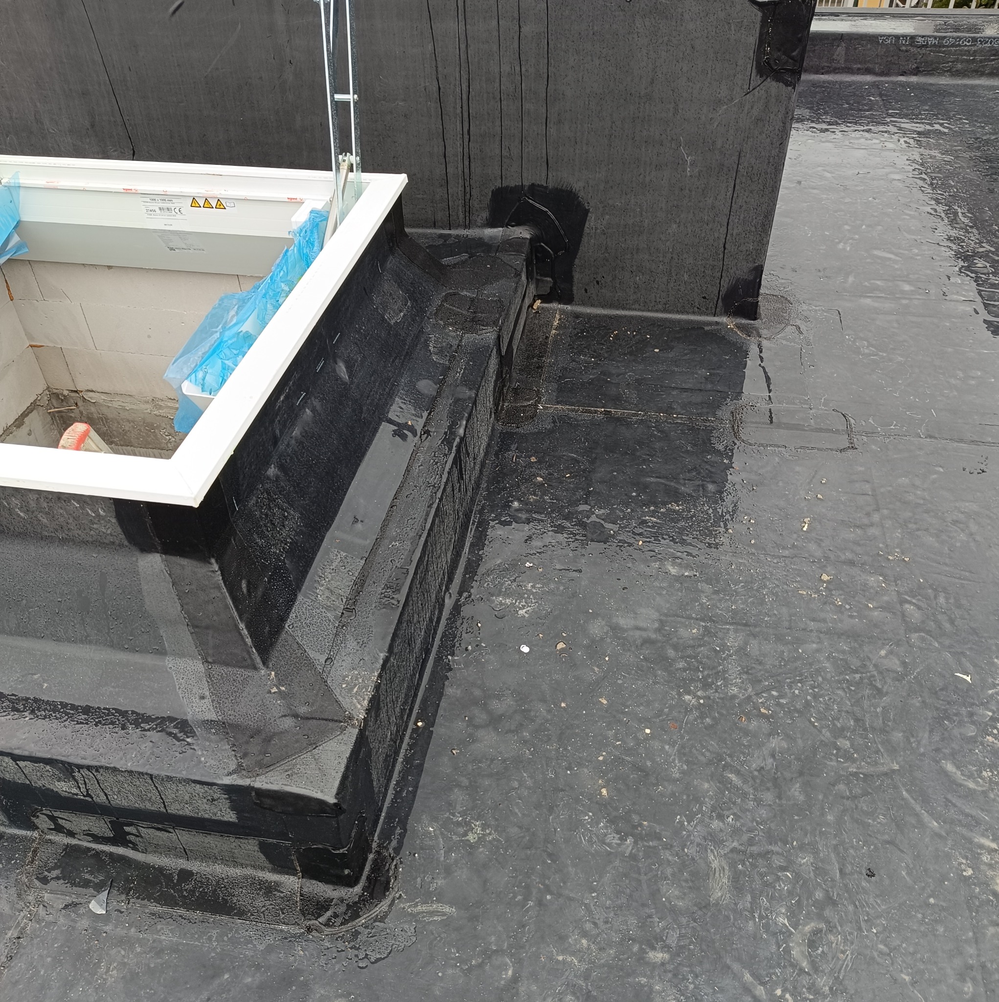 Widok z góry na płaski, czarny dach pokryty papą z zamontowanym świetlikiem dachowym, otoczonym murem z pustaków i zabezpieczonym folią. Na powierzchni dachu widoczne ślady wilgoci i drobne...