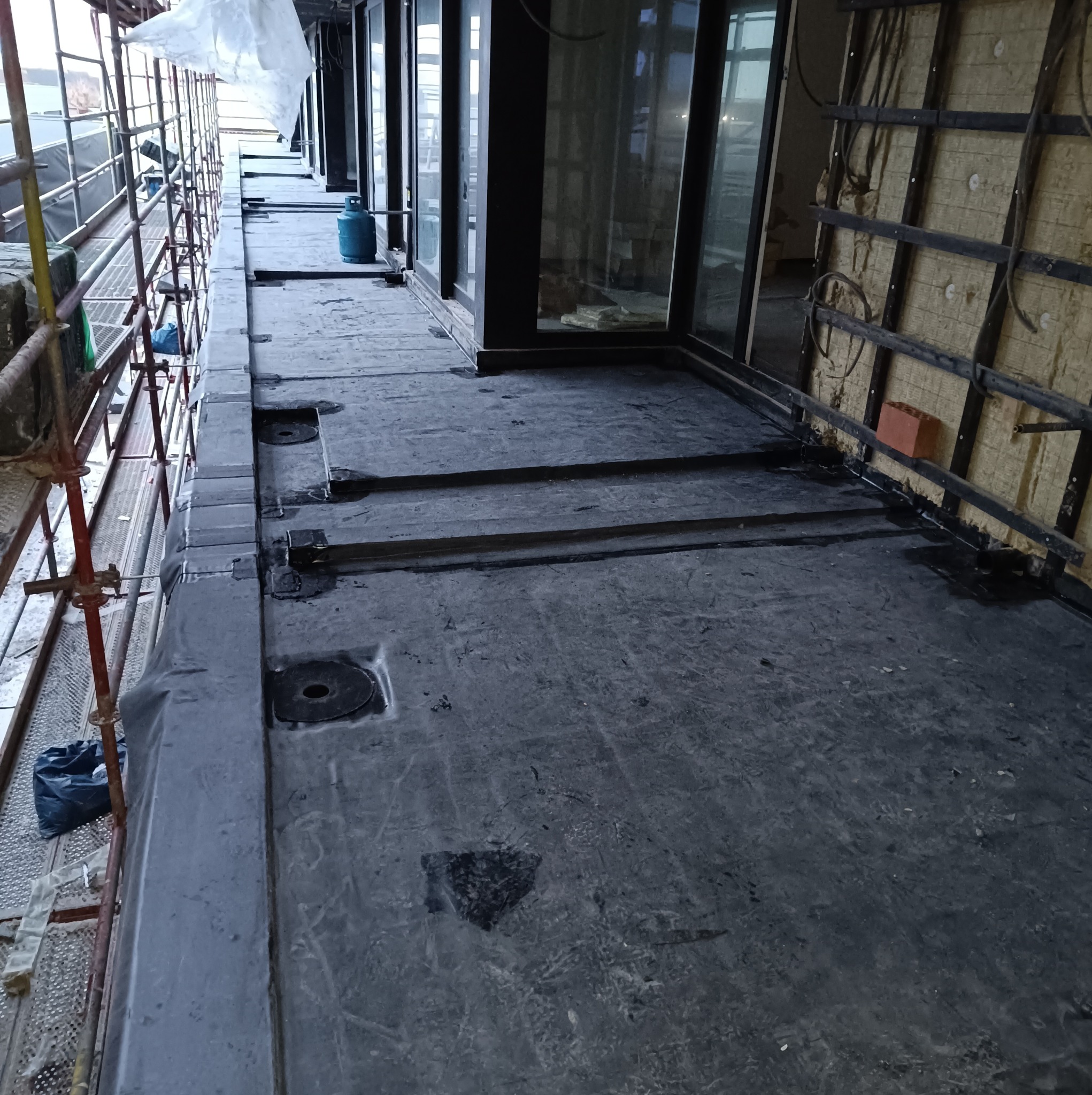 Czarna, płaska powierzchnia z widocznymi okrągłymi otworami i śladami klejenia, prawdopodobnie fragment dachu płaskiego w trakcie prac budowlanych z widocznym rusztowaniem i butlą gazową w tle.