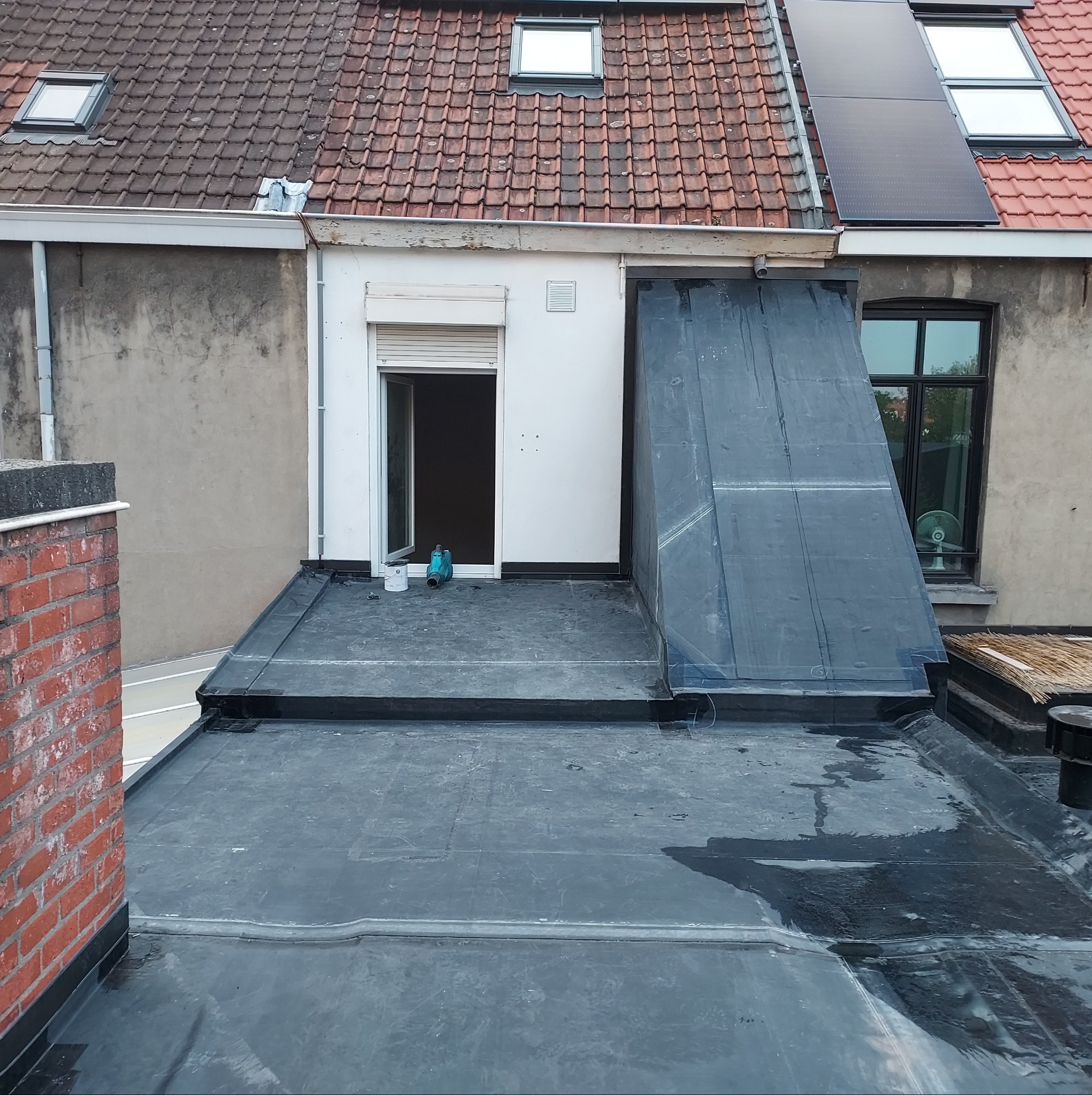 Częściowo pokryty papą płaski dach z widoczną otwartą oknem dachowym, wentylatorem w oknie budynku obok i panelami słonecznymi na dachu budynku w tle.