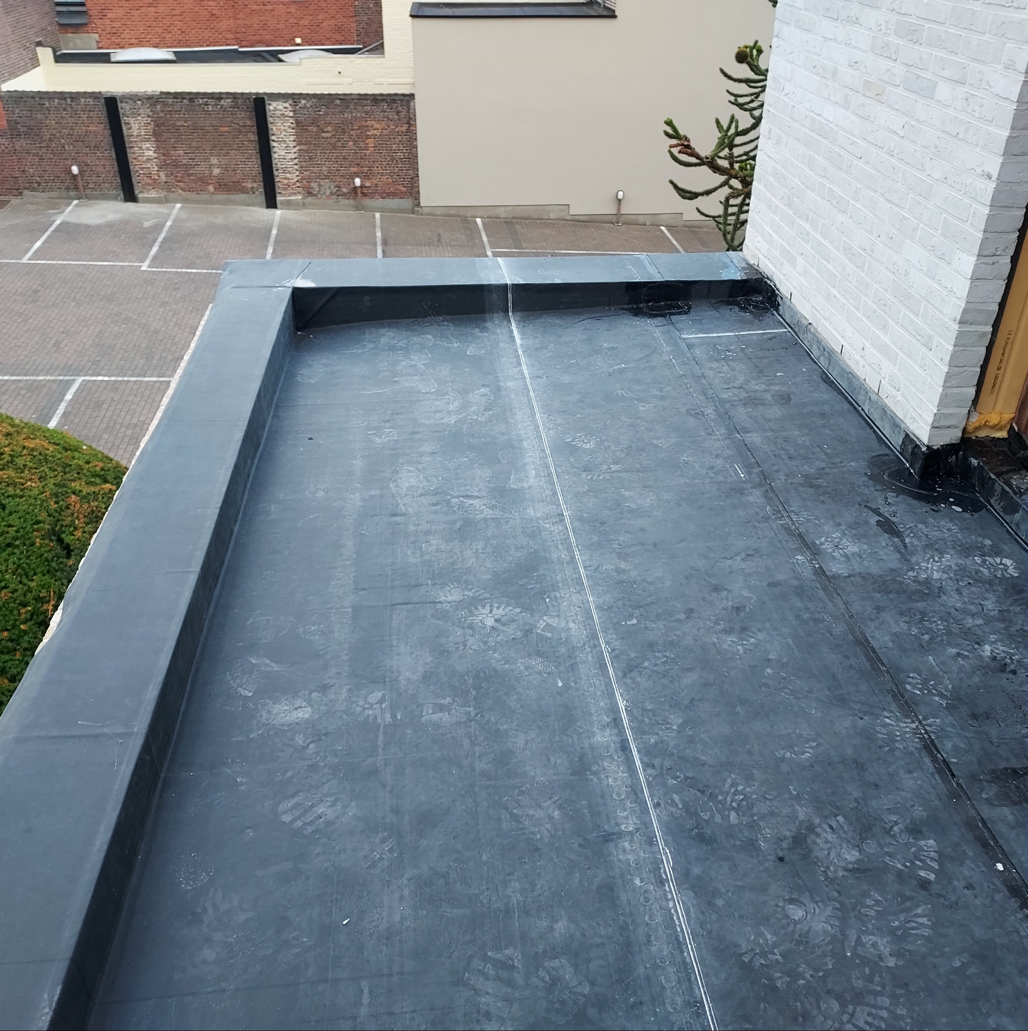 Płaski dach pokryty papą termozgrzewalną, widoczne ślady butów, obróbki blacharskie przy ścianie z białej cegły, w tle budynki i parking.