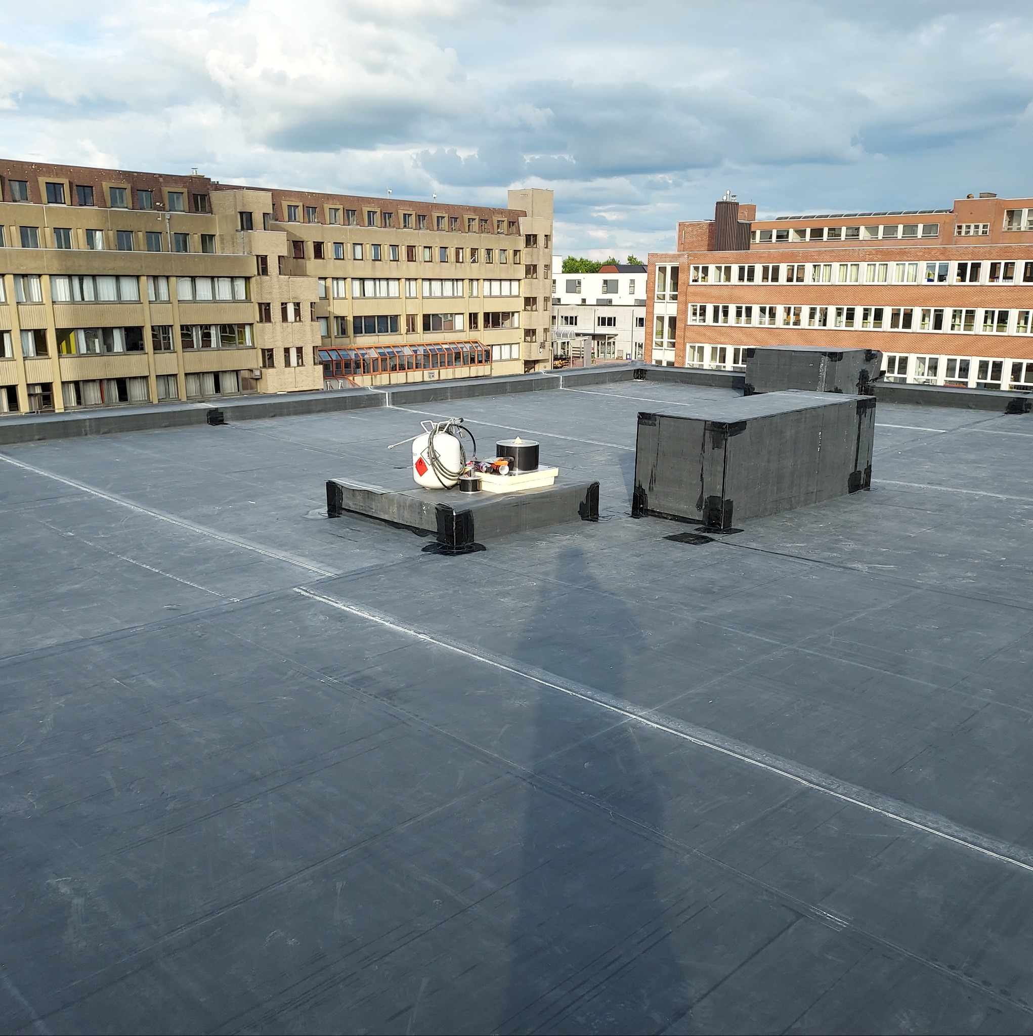 Widok z góry na czarny, płaski dach z papy termozgrzewalnej na tle budynków i pochmurnego nieba; widoczne narzędzia dekarskie i elementy wentylacyjne.