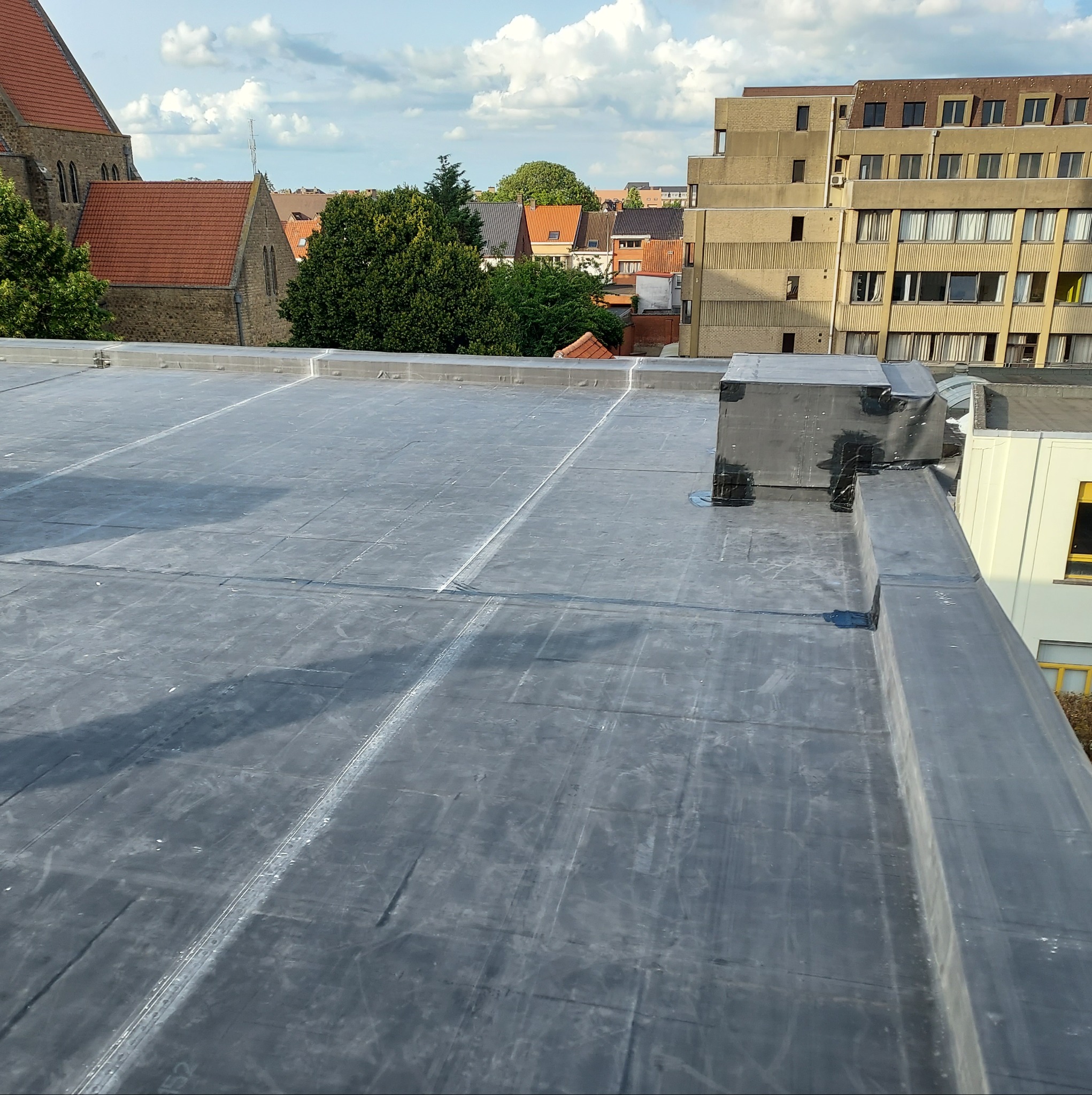 Płaski dach pokryty czarną membraną bitumiczną, widoczne zgrzewy i komin obłożony taśmą izolacyjną, w tle budynki mieszkalne i kościół.