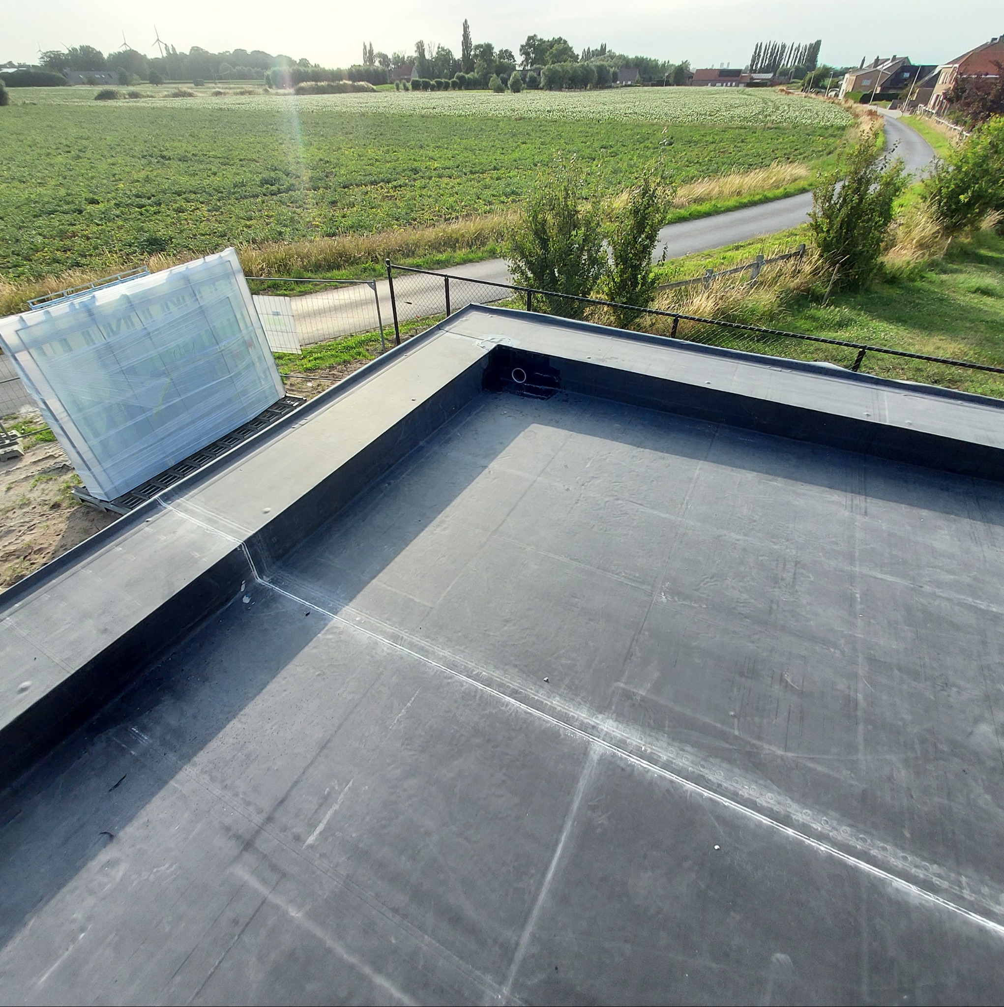 Nowo położona, płaska, czarna papa na dachu z widocznymi łączeniami i odpływem, w tle pole uprawne i zabudowania wiejskie.