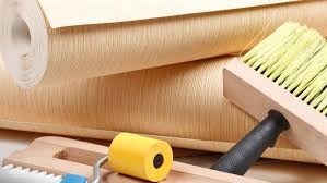 Rolka beżowej tapety o delikatnej fakturze, pędzel do tapetowania z drewnianą rączką i żółty wałek dociskowy na drewnianej podstawie.