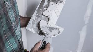 Nakładanie gładzi szpachlowej na ścianę szpachelką, widoczna faktura materiału i fragment ręki w rękawicy ochronnej oraz koszula w kratę osoby wykonującej pracę.