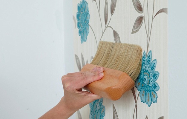 Ręka dociska szczotkę do tapety w kwiatowy wzór, wygładzając powierzchnię na białej ścianie.