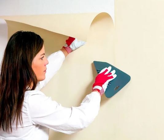 Kobieta w białej koszuli i rękawicach z czerwonymi paskami wygładza ścianę szpachelką, odrywając jednocześnie fragment starej tapety.