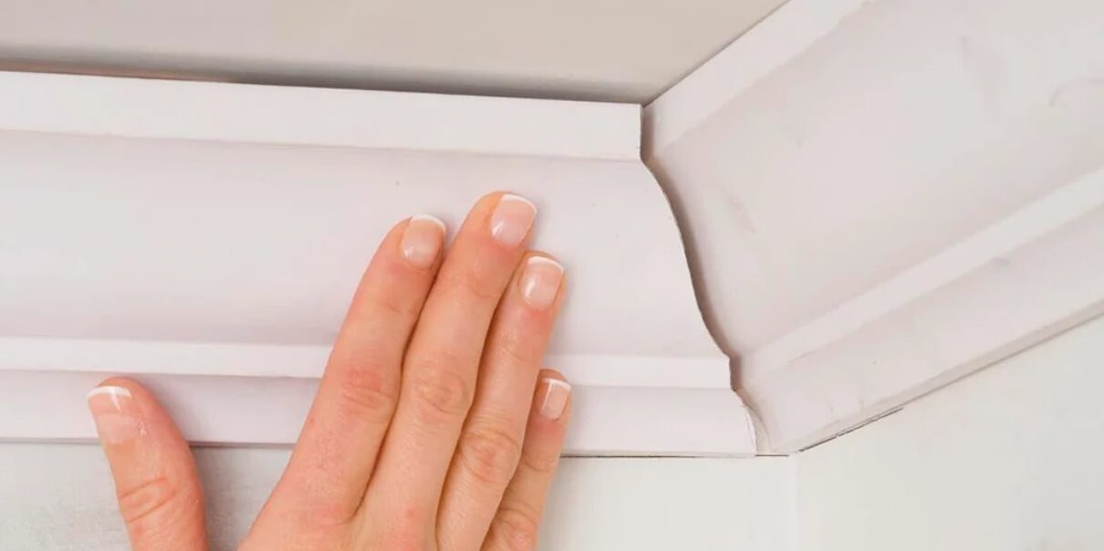 Biała listwa dekoracyjna na styku ściany i sufitu, sprawdzana dotykiem dłoni z francuskim manicure.