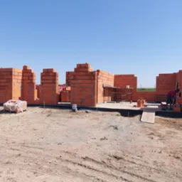 Wznoszenie ścian parterowego domu z czerwonej cegły w Mogilnie, widoczne fragmenty ścian, paleta cegieł w folii, pracownik przy pracy, w tle turbiny wiatrowe.