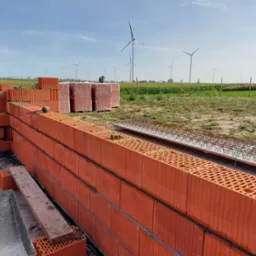 Budowa domu z czerwonej cegły ceramicznej Porotherm na fundamencie, w tle pole uprawne i turbiny wiatrowe.