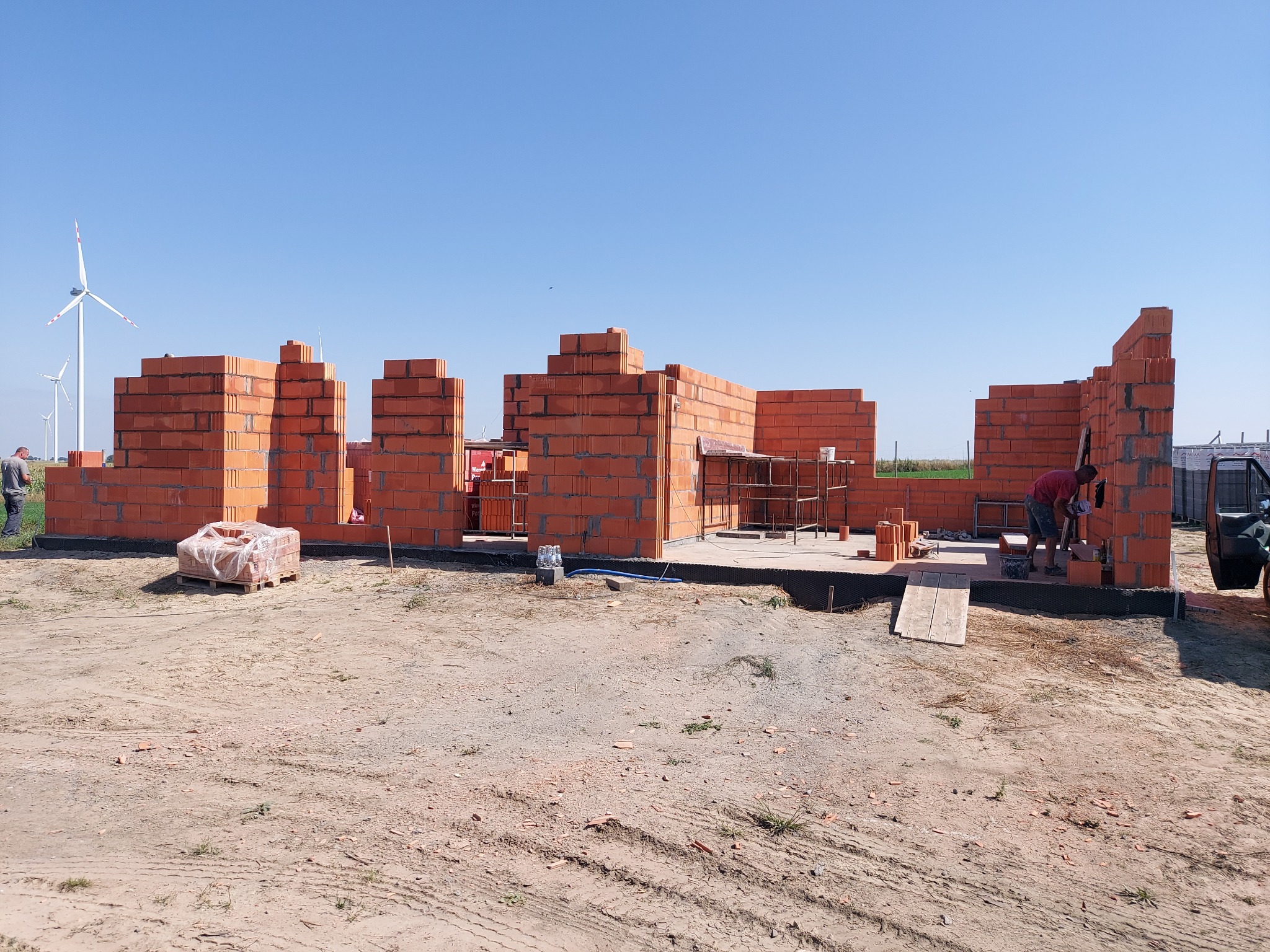 Wznoszenie ścian parterowego domu z czerwonej cegły w Mogilnie, widoczne fragmenty ścian, paleta cegieł w folii, pracownik przy pracy, w tle turbiny wiatrowe.