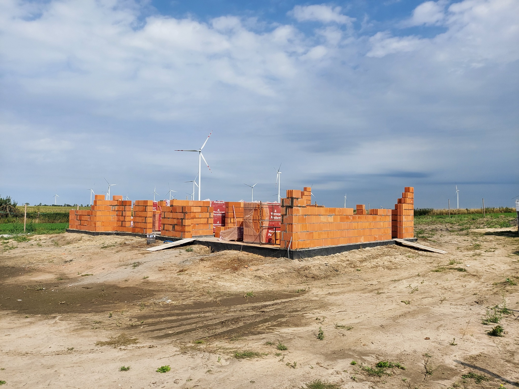 Wznoszone mury parterowego domu z czerwonej cegły na fundamencie, w tle turbiny wiatrowe pod pochmurnym niebem.