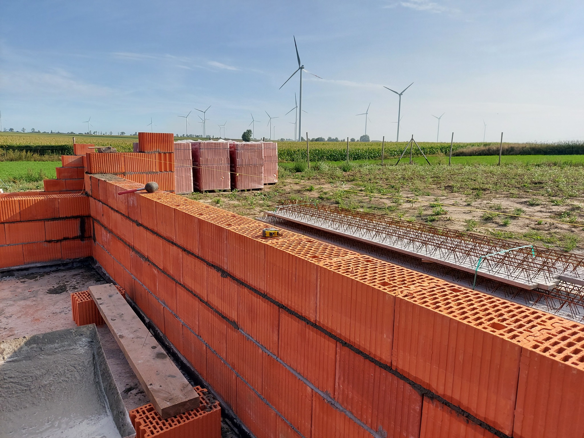 Budowa domu z czerwonej cegły ceramicznej Porotherm na fundamencie, w tle pole uprawne i turbiny wiatrowe.