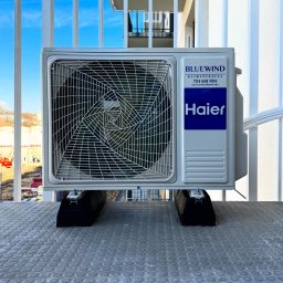 Blue Wind Klimatyzacja - Biała jednostka zewnętrzna klimatyzacji Haier zamontowana na balkonie, widoczne logo Bluewind i dane kontaktowe firmy. Tło: błękitne niebo i fragment budynku.