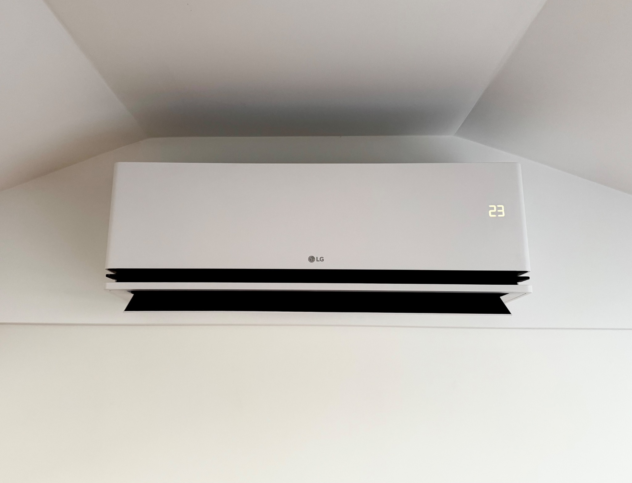 Biała klimatyzacja LG na białej ścianie pod skośnym sufitem, wyświetlająca temperaturę 23 stopnie.