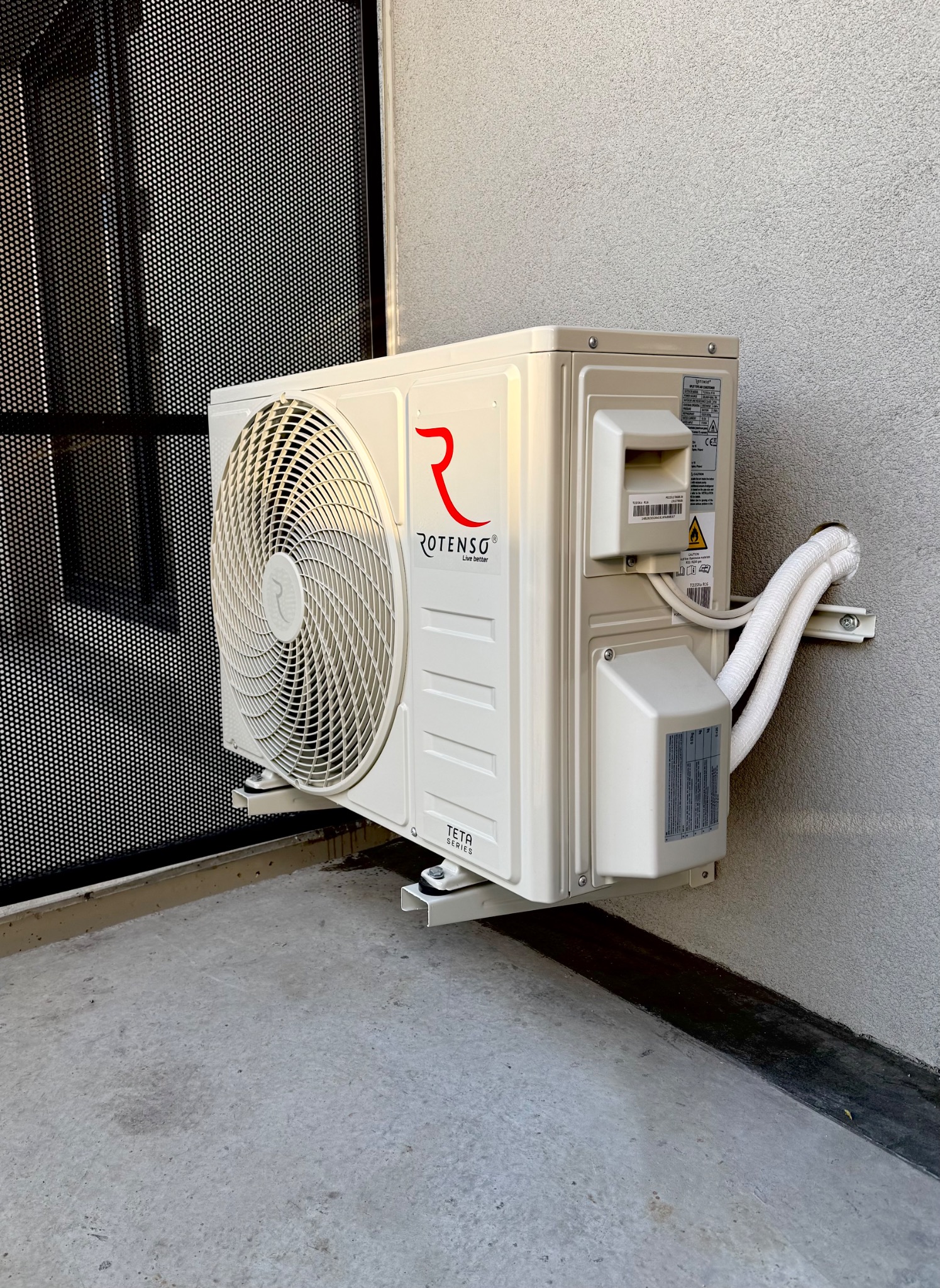 Zewnętrzna jednostka klimatyzacji Rotenso Teta Series zamontowana na jasnoszarym budynku z widocznymi przewodami i kratką wentylacyjną w tle.