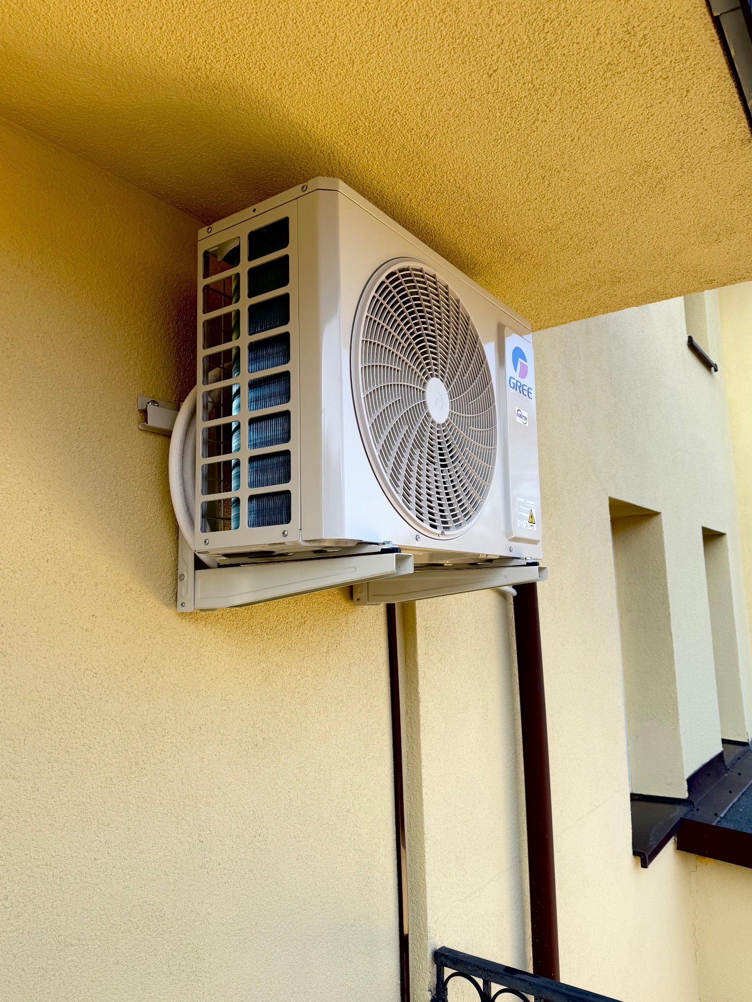 Zewnętrzna jednostka klimatyzatora marki Gree zamontowana na żółtej, teksturowanej ścianie budynku, widoczna z dołu pod kątem, z elementami instalacji i logo producenta.