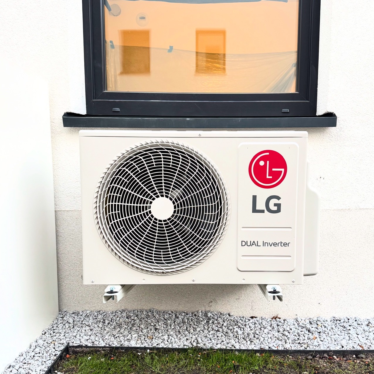 Zamontowana jednostka zewnętrzna klimatyzatora LG Dual Inverter na elewacji budynku, widok z dołu, szare kamienie i trawa u podstawy ściany.