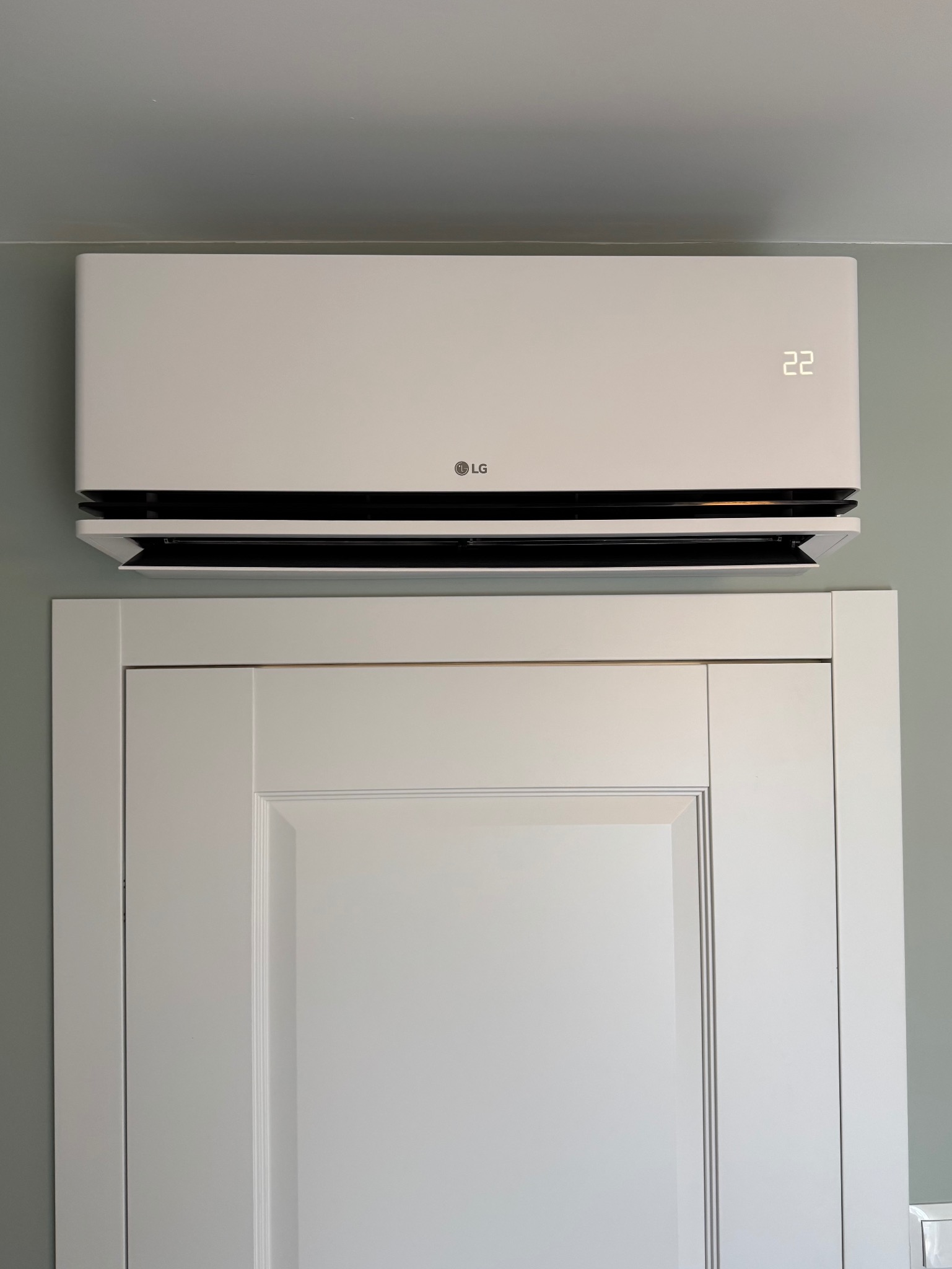 Biała jednostka wewnętrzna klimatyzacji LG zamontowana nad białymi drzwiami w pomieszczeniu o ścianach w kolorze gołębim.