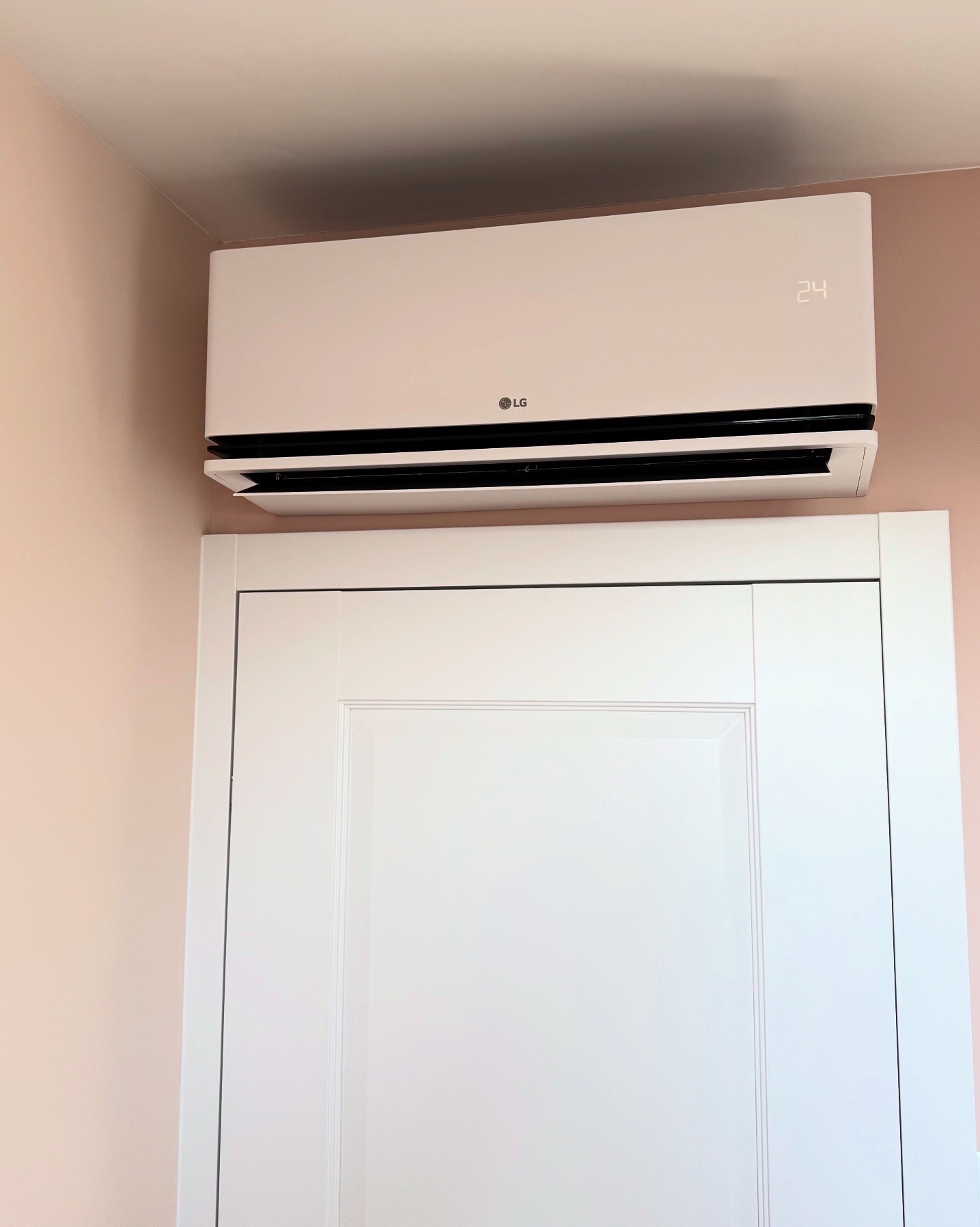 Biały klimatyzator LG z wyświetlaczem temperatury 24 stopnie Celsjusza, zamontowany nad białymi drzwiami w pomieszczeniu o ścianach w kolorze pudrowego różu.