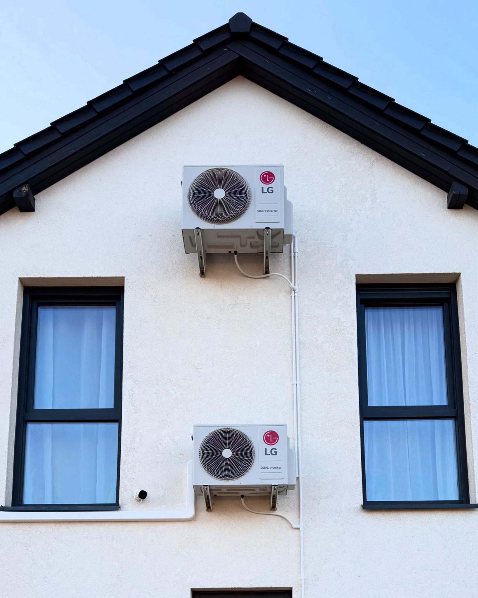 Dwa moduły zewnętrzne klimatyzacji LG Dual Inverter zamontowane na białej fasadzie domu z czarnym dachem i ramami okiennymi.