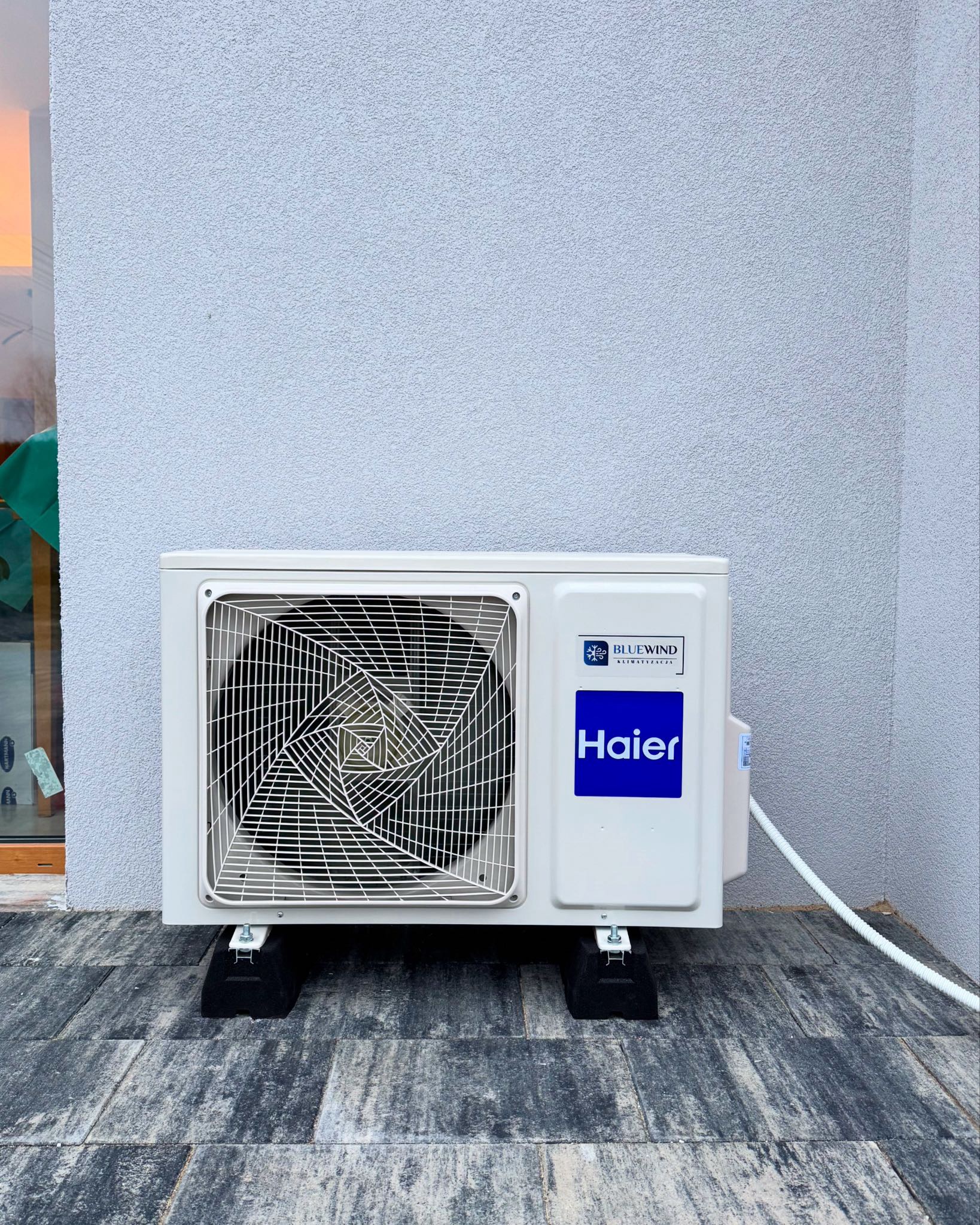 Zewnętrzna jednostka klimatyzacji Haier z logo BlueWind, zamontowana na czarnych podkładkach na szarym brukowanym patio, na tle teksturowanej ściany budynku.