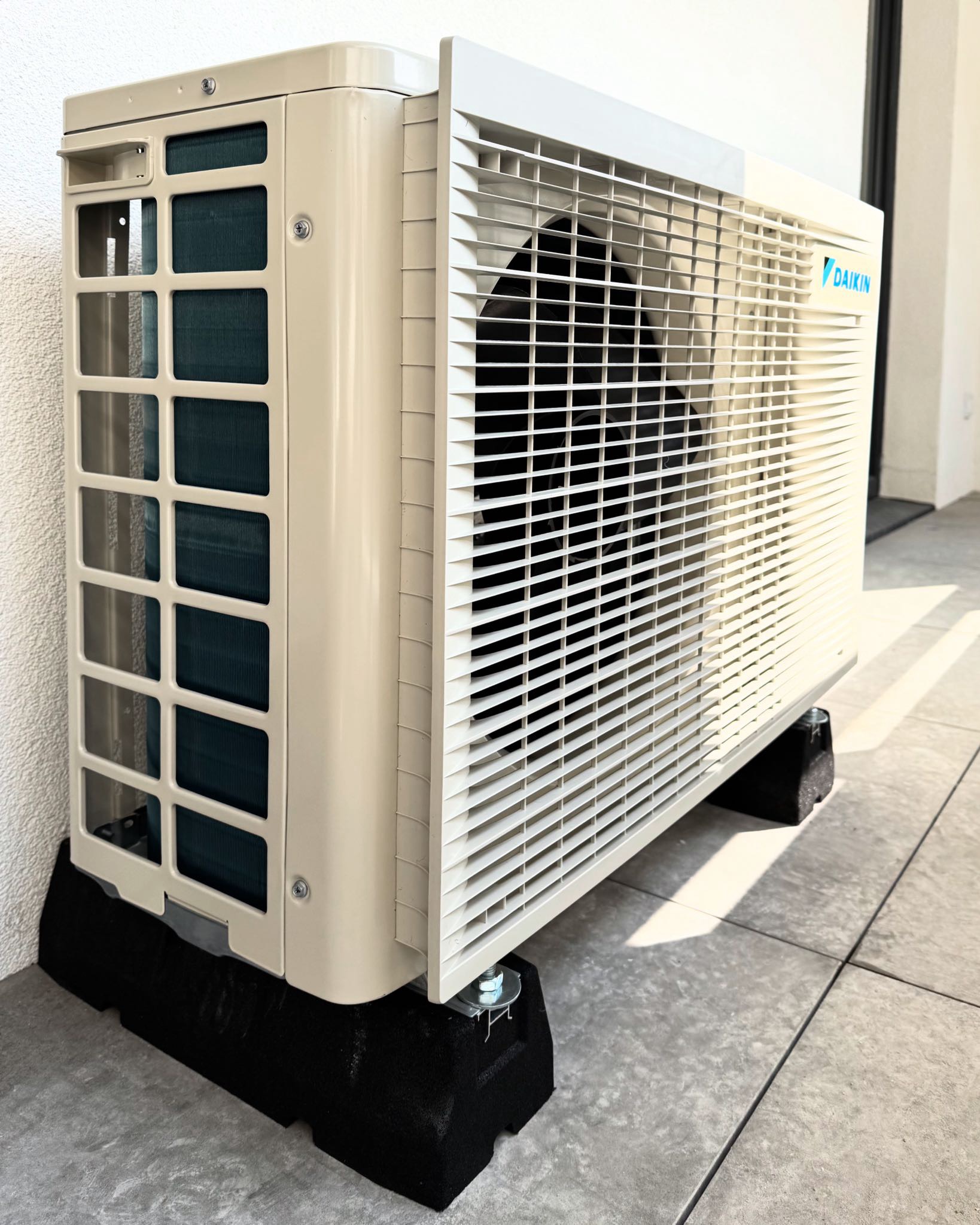 Zewnętrzna jednostka klimatyzatora Daikin zamontowana na czarnych, antywibracyjnych podkładkach na jasnoszarym, wyłożonym płytkami tarasie przy białej ścianie budynku.