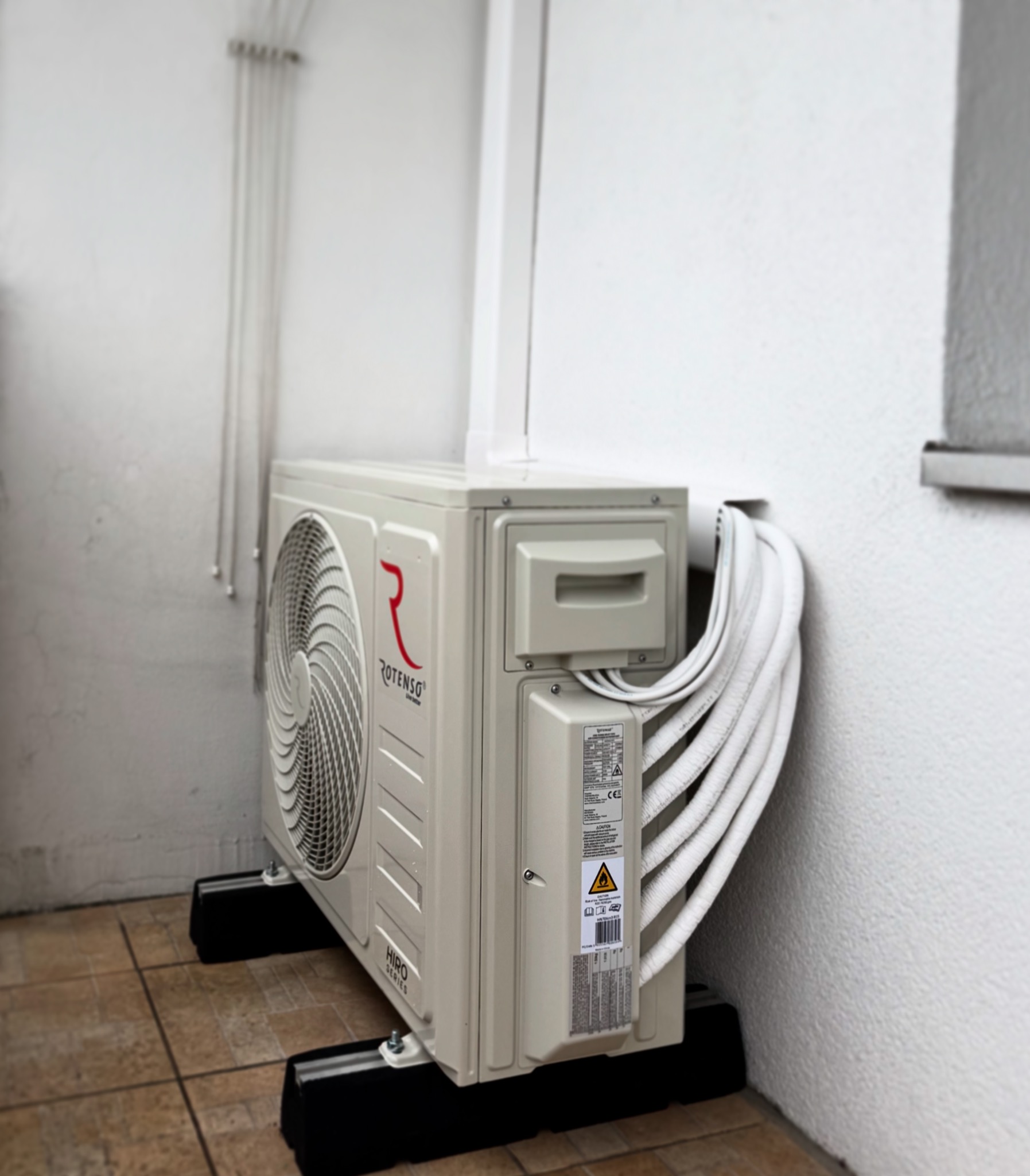Zewnętrzna jednostka klimatyzatora Rotenso Hiro Series zamontowana na czarnych podstawkach na brązowej, kafelkowej podłodze, z widocznymi rurami prowadzącymi do wnętrza budynku.