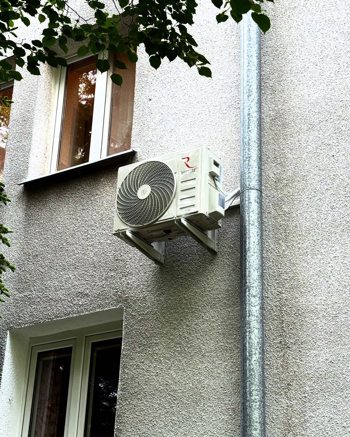 Biały klimatyzator na szarej, teksturowanej ścianie budynku, widoczny obok okna i rury spustowej. Zewnętrzna jednostka klimatyzacji zamontowana na wspornikach.