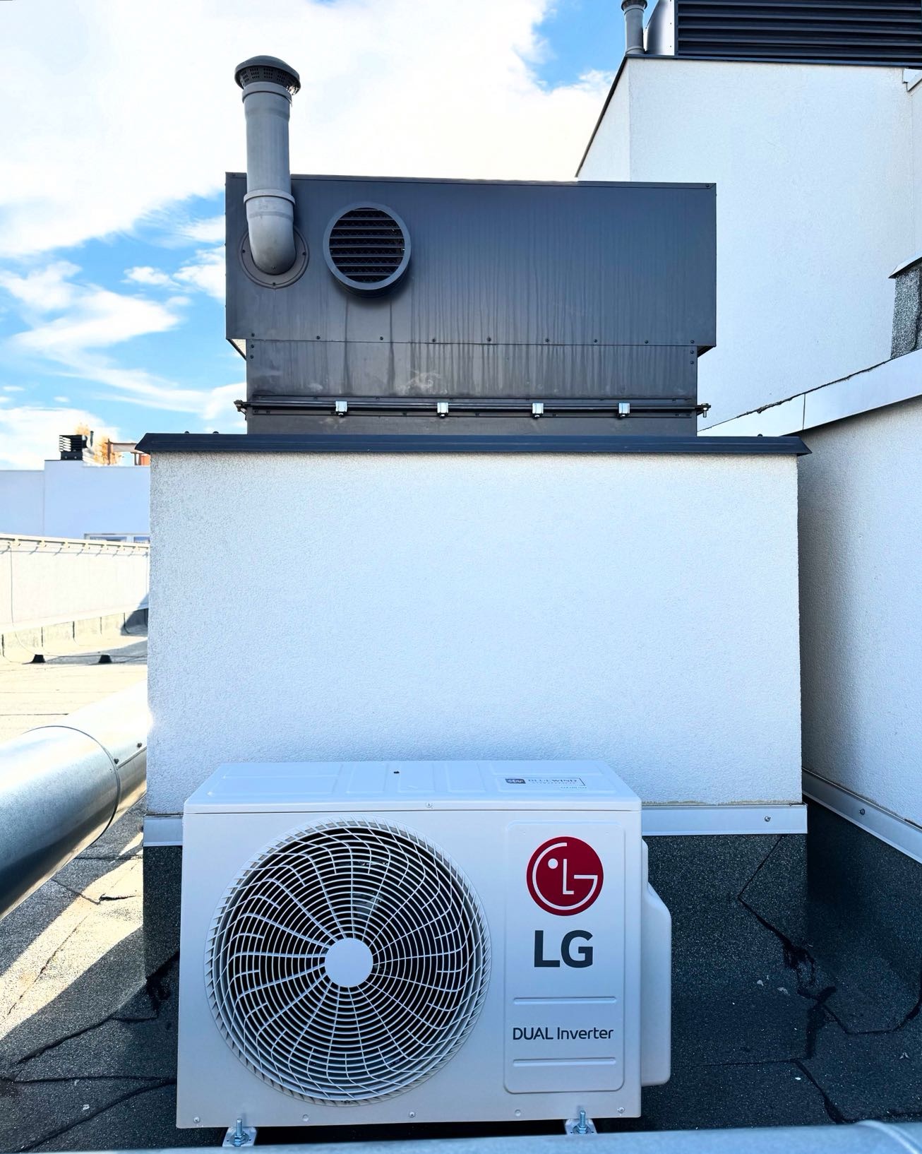 Biała jednostka zewnętrzna klimatyzacji LG Dual Inverter na dachu budynku, z widoczną instalacją wentylacyjną i błękitnym niebem w tle. Lekki, słoneczny dzień.