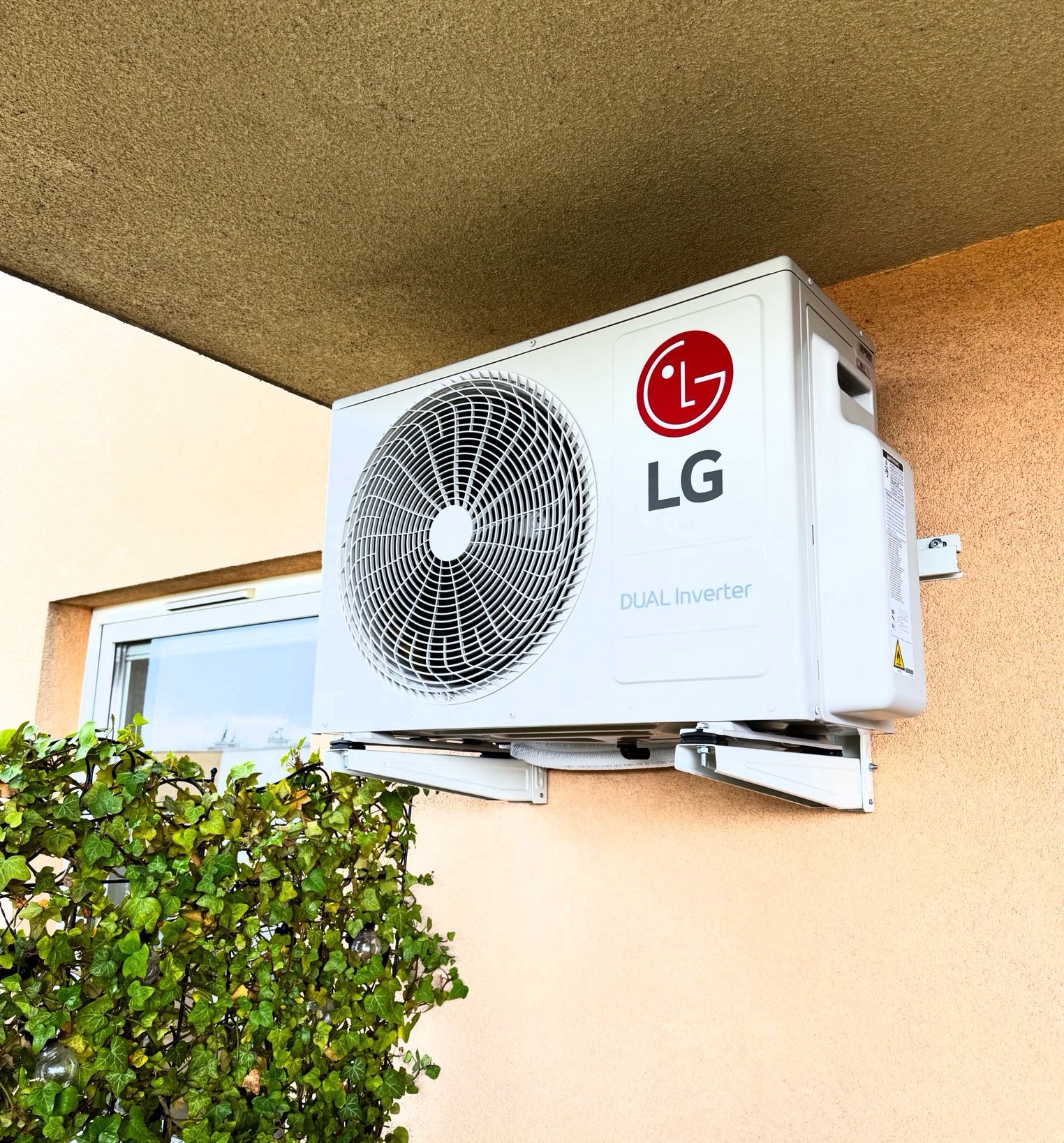Biała jednostka zewnętrzna klimatyzacji LG Dual Inverter zamontowana na ścianie budynku, z zielonymi roślinami w tle i oknem po lewej stronie.