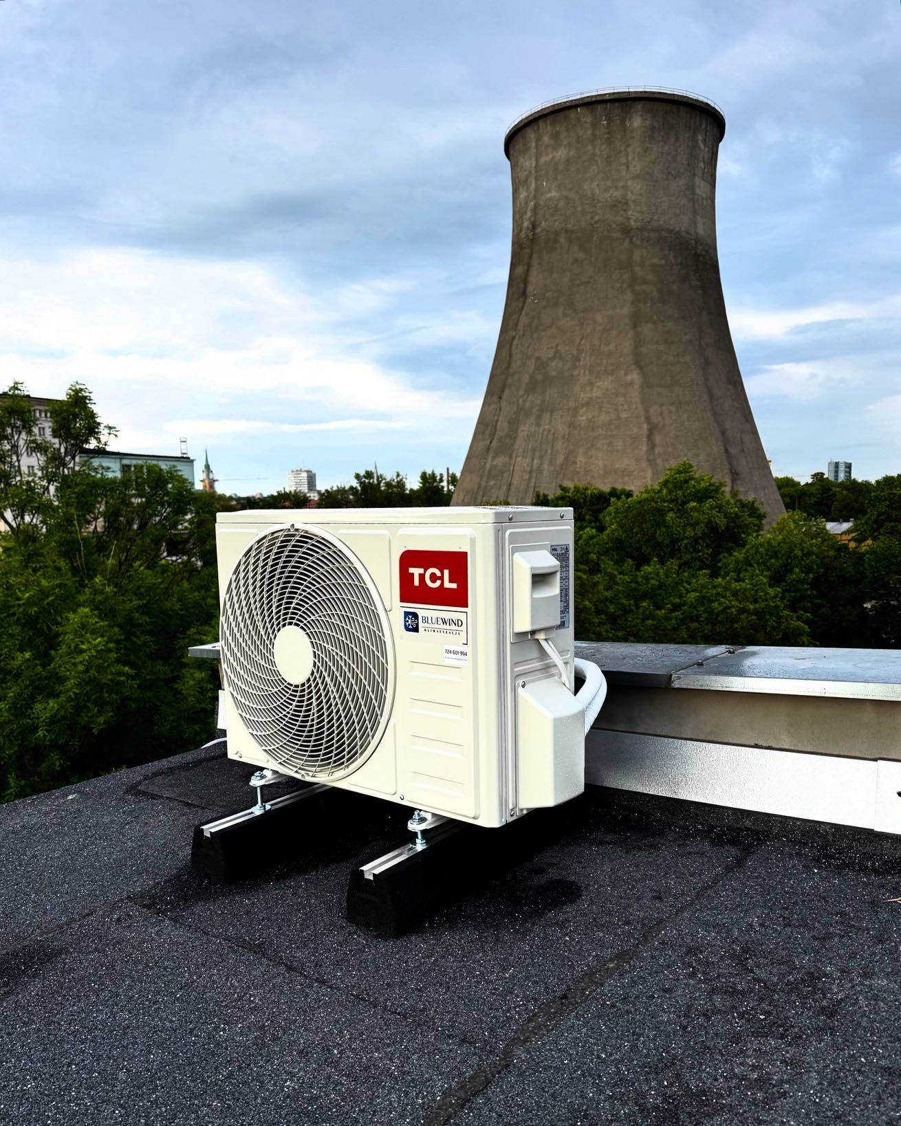 Klimatyzator TCL zamontowany na dachu budynku w Łodzi, z charakterystyczną chłodnią kominową w tle. Widok na miasto i zieleń. Instalacja klimatyzacji.