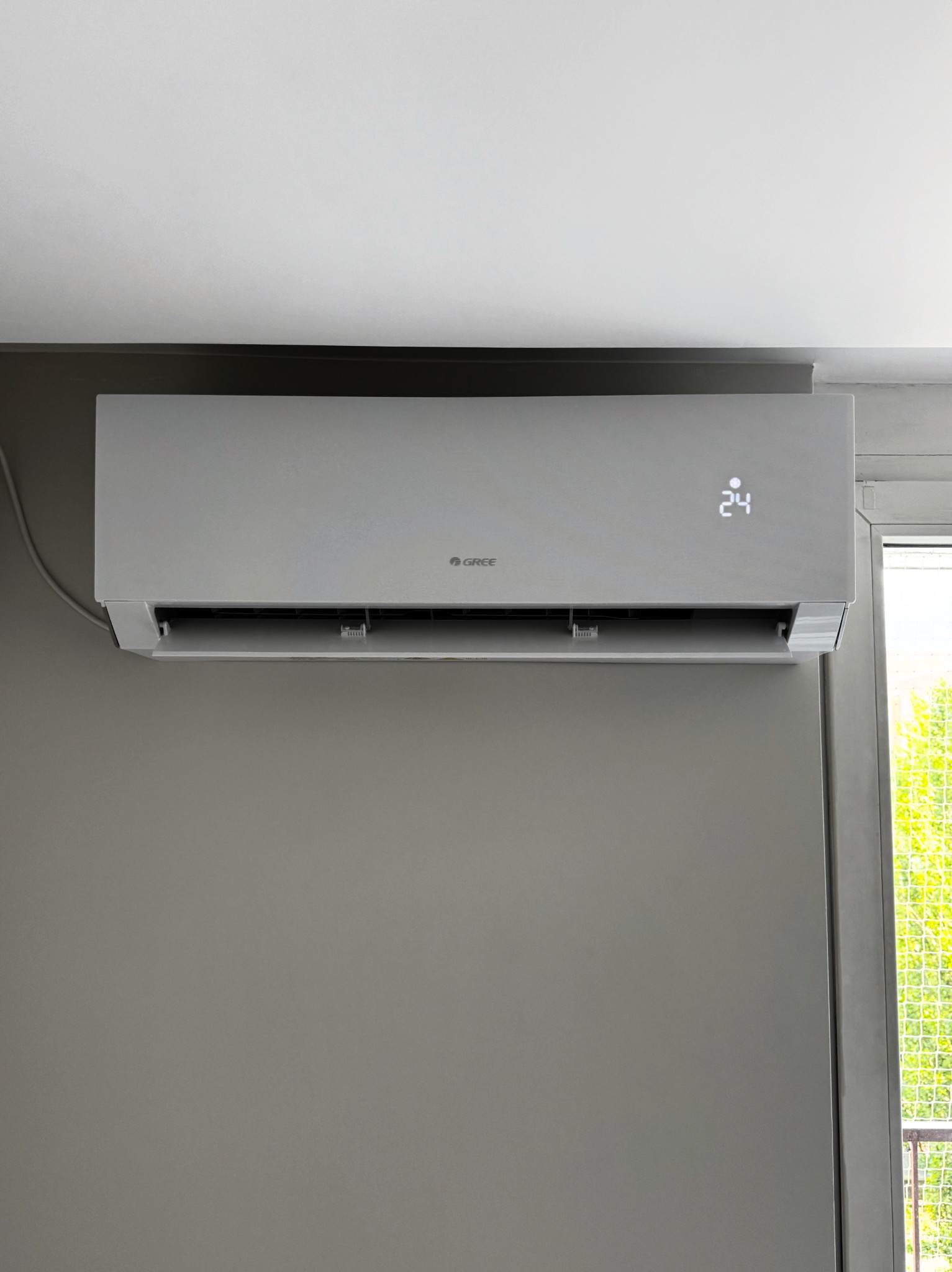 Szary klimatyzator Gree z wyświetlaczem 24 stopnie Celsjusza, zamontowany na szarej ścianie, obok okna z widokiem na zieleń. Kabel zasilający widoczny po lewej.