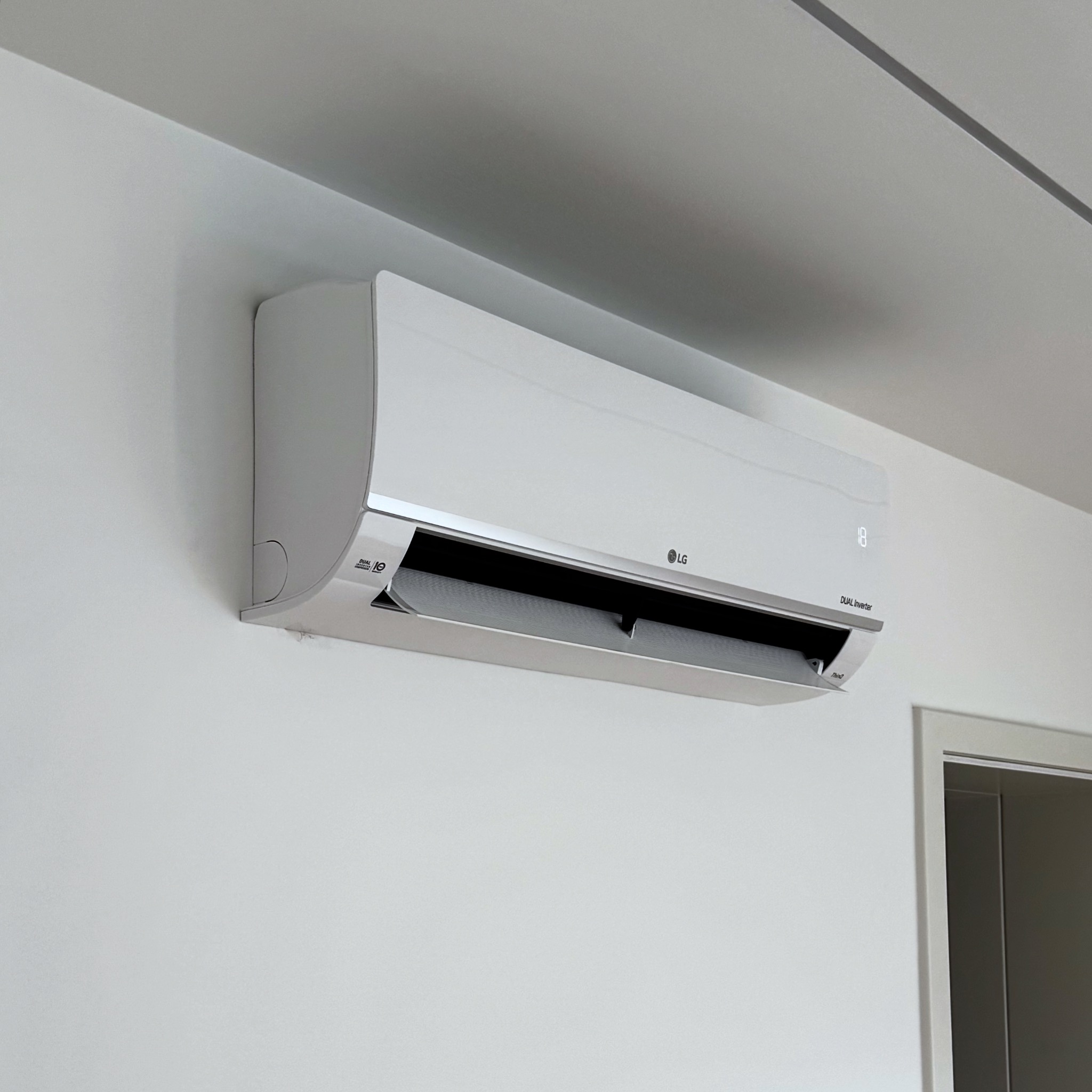 Biała klimatyzacja LG Dual Inverter zamontowana na białej ścianie w jasnym pomieszczeniu. Widoczny wyświetlacz temperatury i fragment drzwi.
