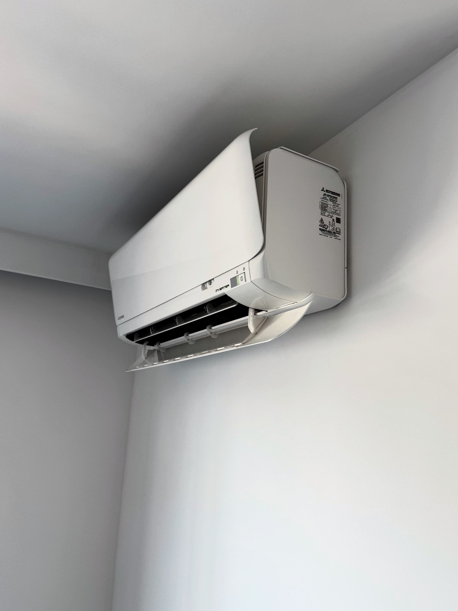 Biała klimatyzacja Mitsubishi Inverter zamontowana na ścianie w rogu jasnego pomieszczenia. Widok z dołu, lekki kąt.