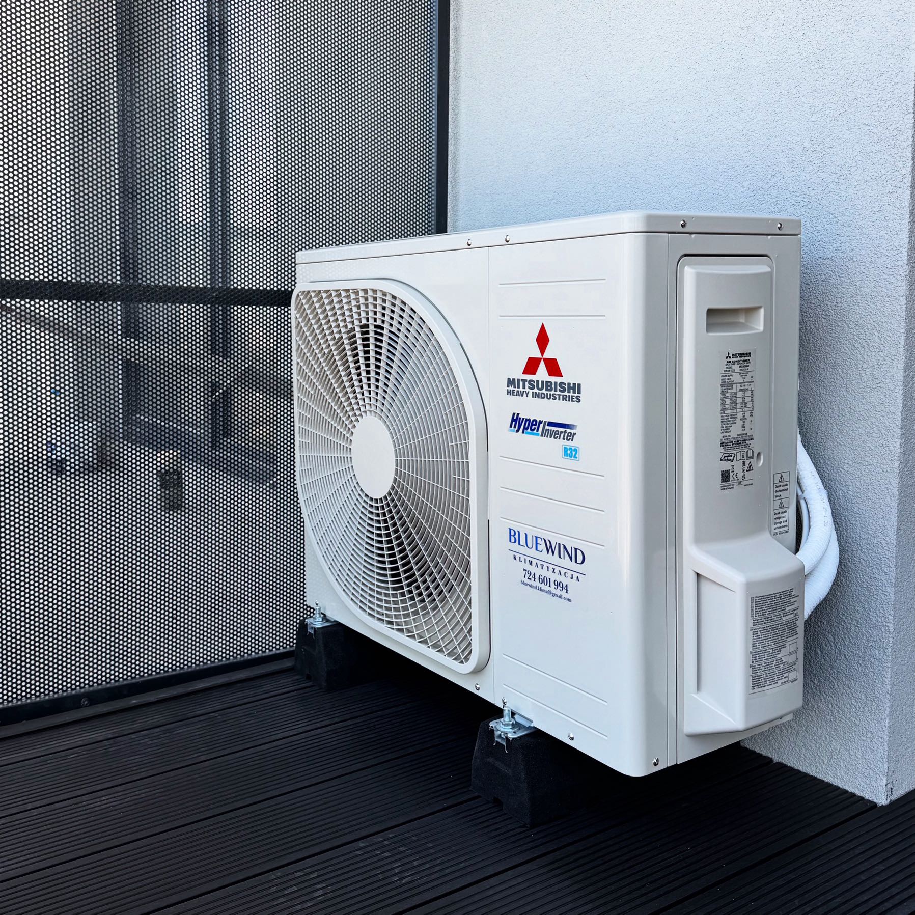 Biała jednostka zewnętrzna klimatyzacji Mitsubishi Heavy Industries, model Hyper Inverter R32, zamontowana na balkonie z czarną podłogą i ażurową balustradą.