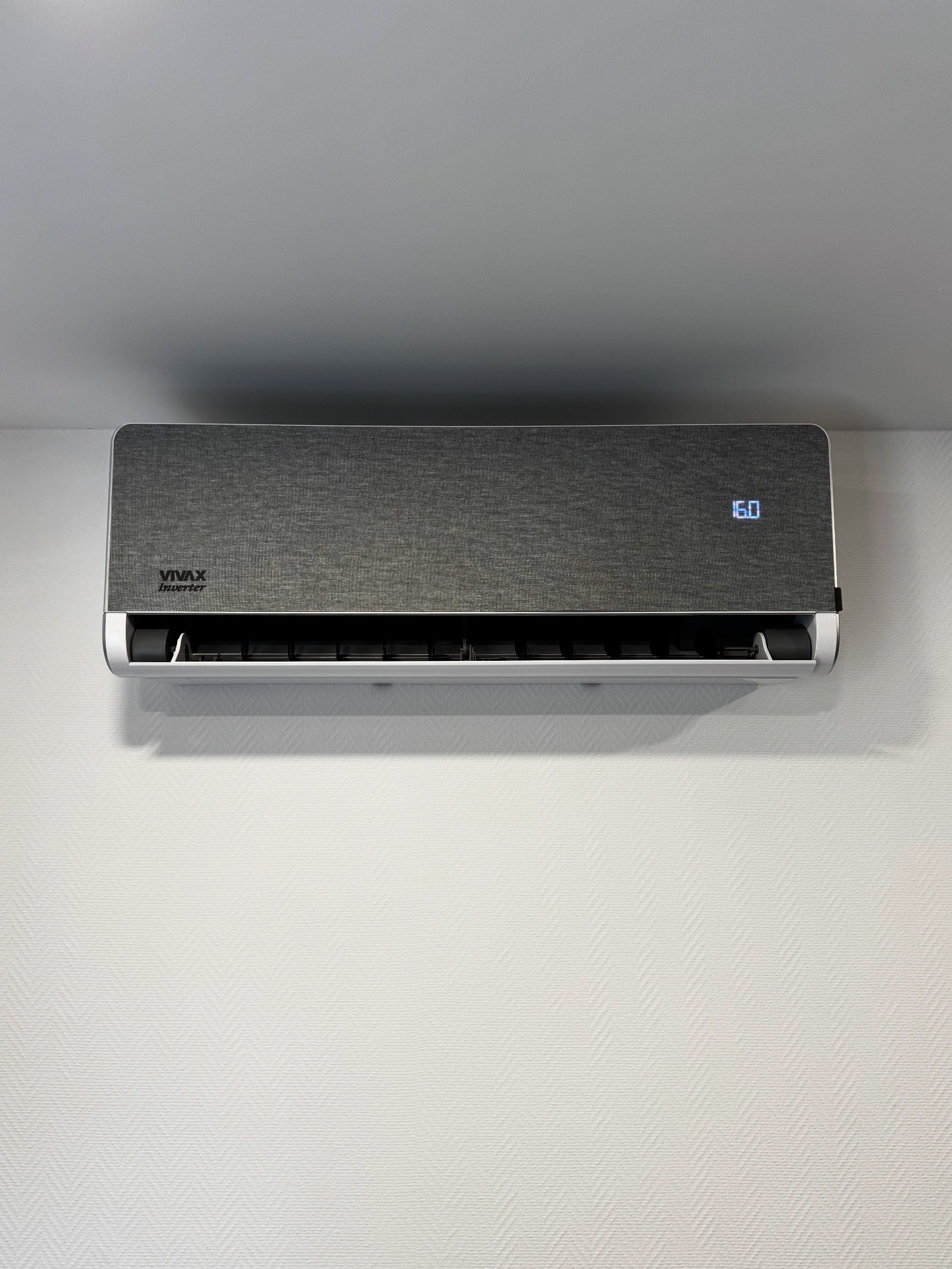 Szary klimatyzator VIVAX Inverter z wyświetlaczem 16.0, zamontowany na białej ścianie z delikatną fakturą. Widok z przodu, oświetlenie naturalne.