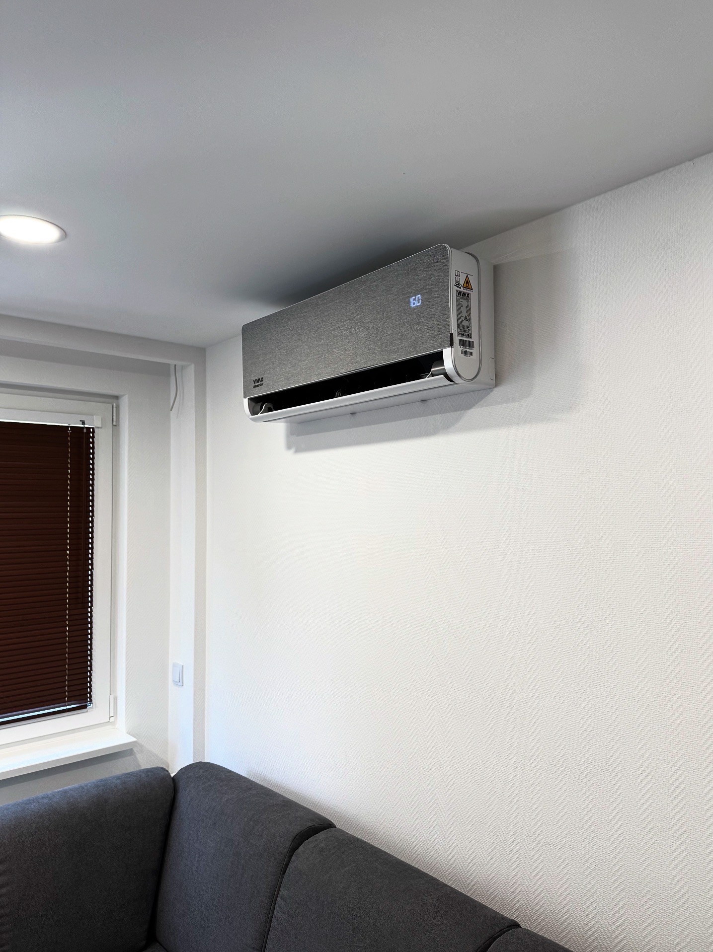 Nowoczesny klimatyzator ścienny w szarym odcieniu, zamontowany w jasnym pokoju z szarą kanapą i oknem z żaluzjami. Wyświetlacz pokazuje temperaturę 16 stopni.