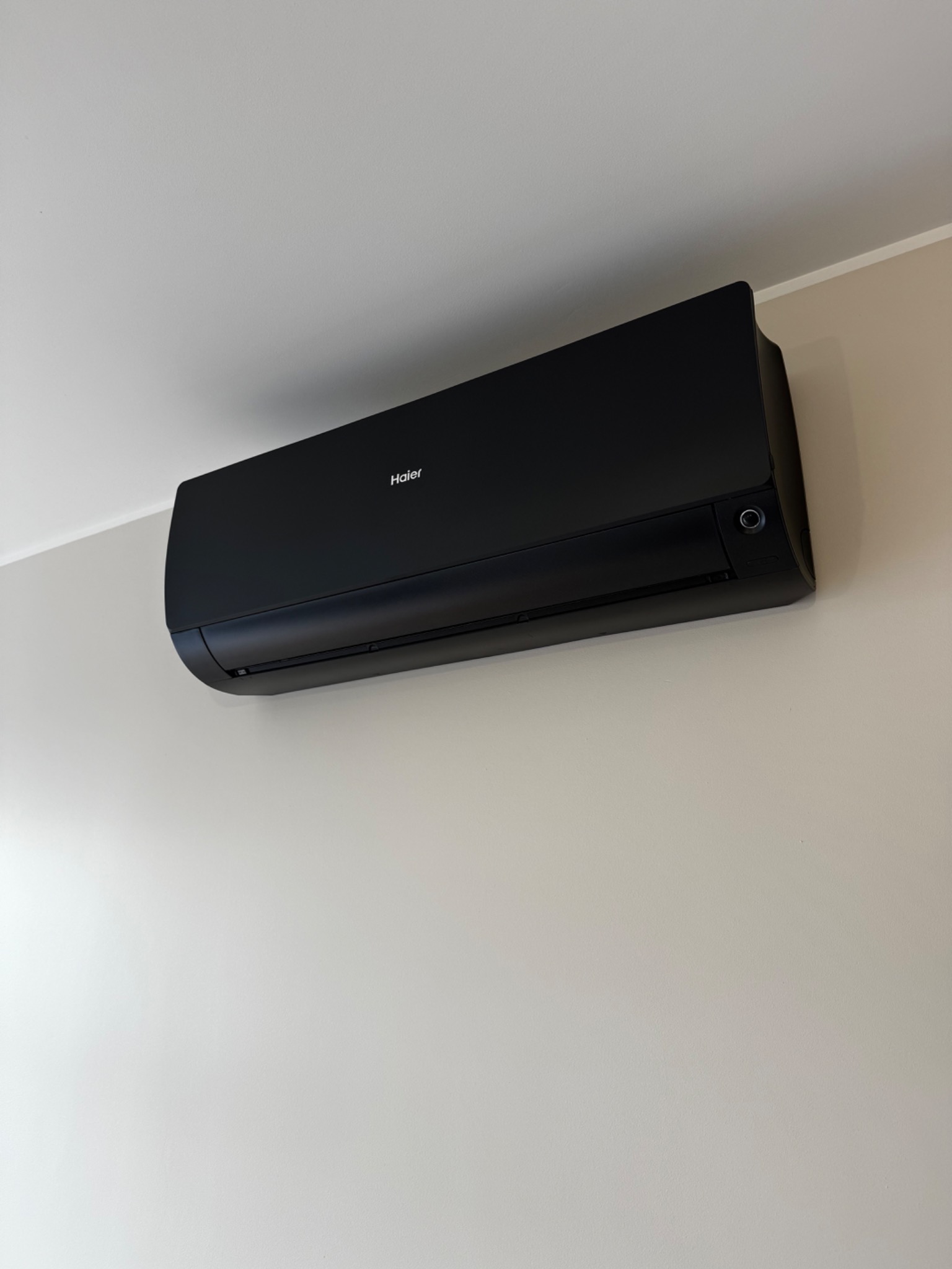 Czarny klimatyzator Haier zamontowany na jasnej ścianie. Widoczny z boku, z delikatnym oświetleniem, tworzy nowoczesny wygląd.