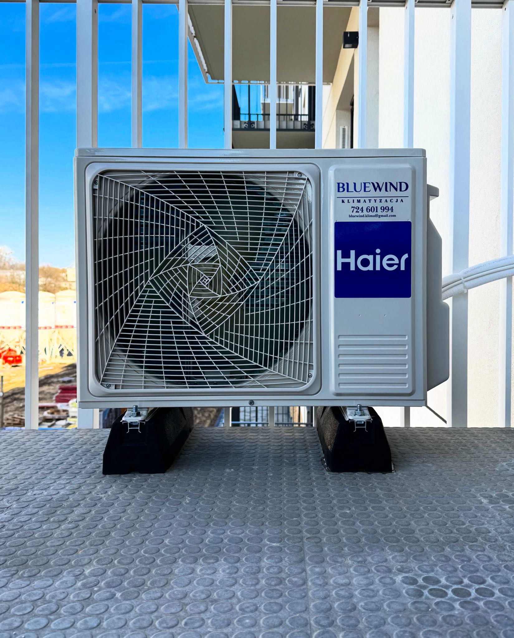 Biała jednostka zewnętrzna klimatyzacji Haier zamontowana na balkonie, widoczne logo Bluewind i dane kontaktowe firmy. Tło: błękitne niebo i fragment budynku.