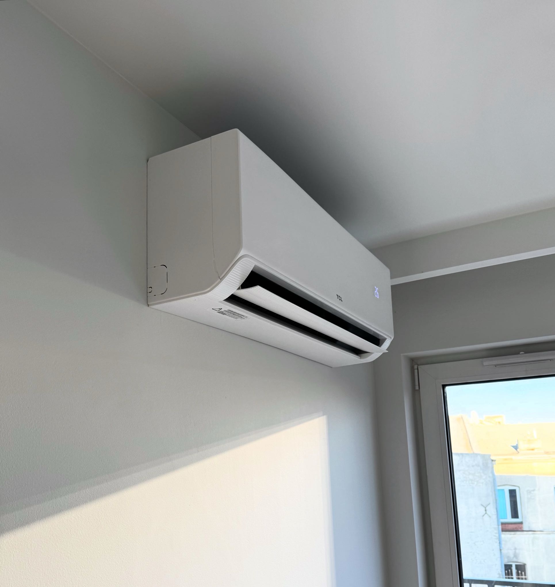 Biały klimatyzator TCL zamontowany na ścianie w jasnym pomieszczeniu, widoczne okno z widokiem na budynek. Delikatne światło słoneczne podkreśla czystość wnętrza.