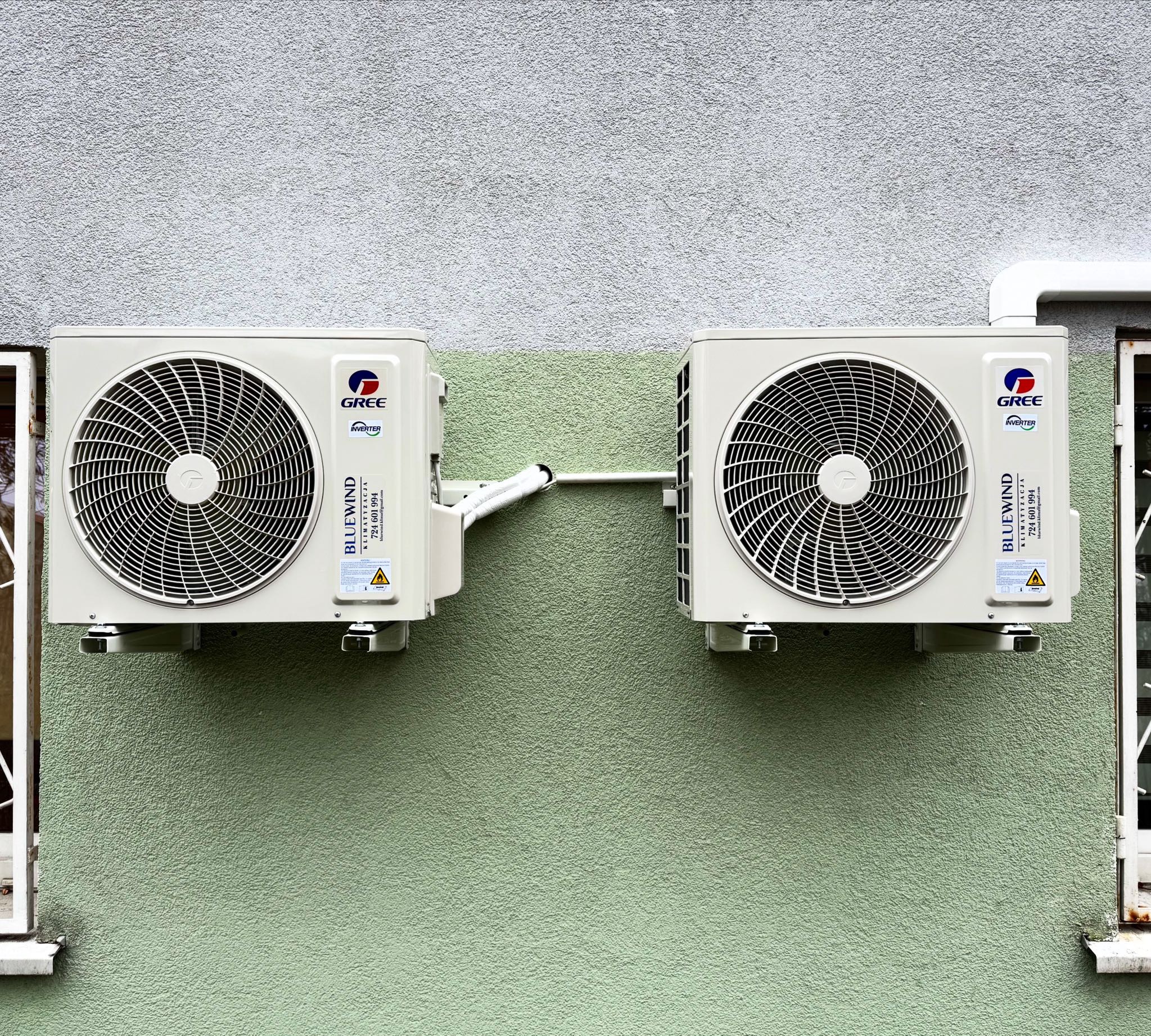 Dwa białe klimatyzatory Gree BlueWind zamontowane na zielonej ścianie budynku, połączone białymi rurami. Widoczne fragmenty okien po bokach.
