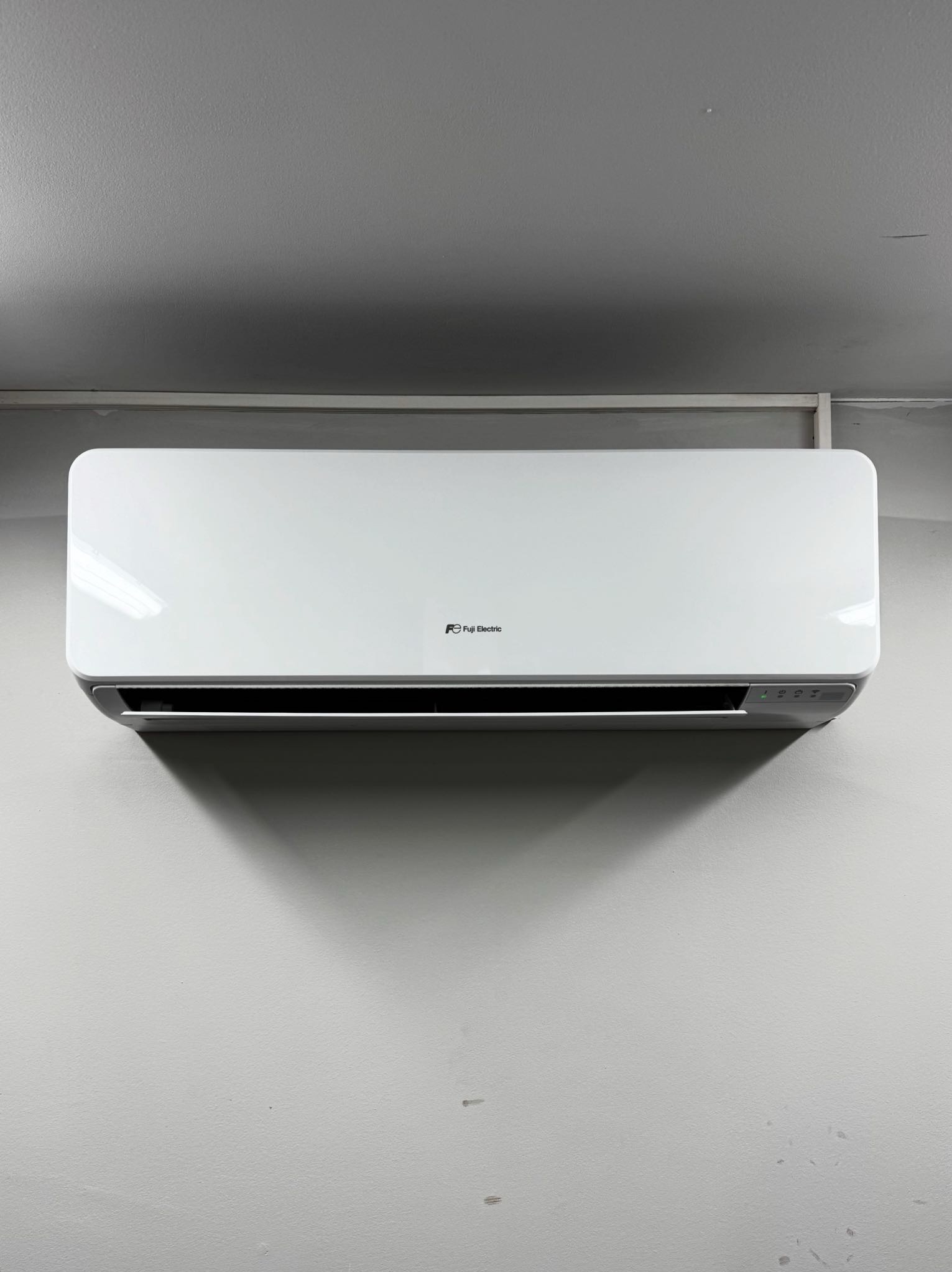 Biała klimatyzacja Fuji Electric zamontowana na szarej ścianie. Widoczne diody sygnalizacyjne na urządzeniu. Minimalistyczny design.