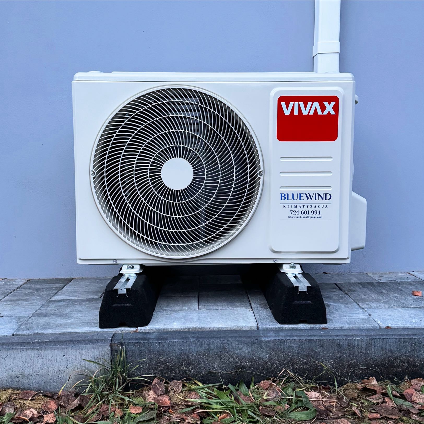 Biała jednostka zewnętrzna klimatyzacji VIVAX na czarnych podkładkach, zamontowana przy jasnoniebieskiej ścianie budynku. Widoczne logo BlueWind i dane kontaktowe.