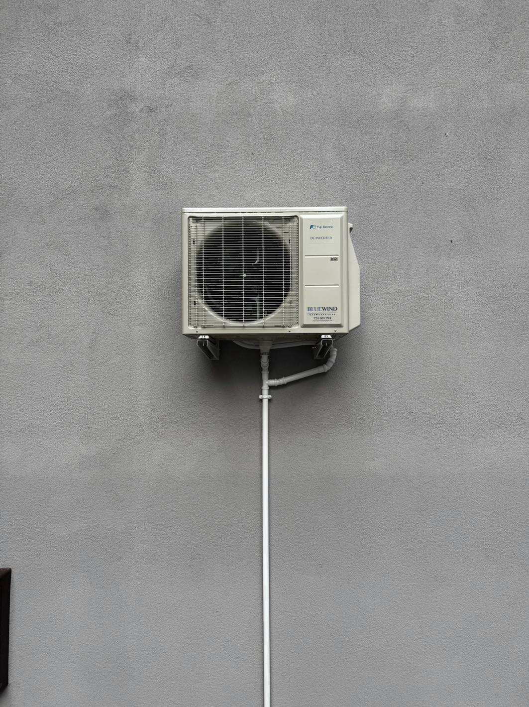 Zamontowana jednostka zewnętrzna klimatyzacji Fuji Electric BlueWind na szarej ścianie budynku. Widoczne rury odprowadzające skropliny. Minimalistyczny kadr.